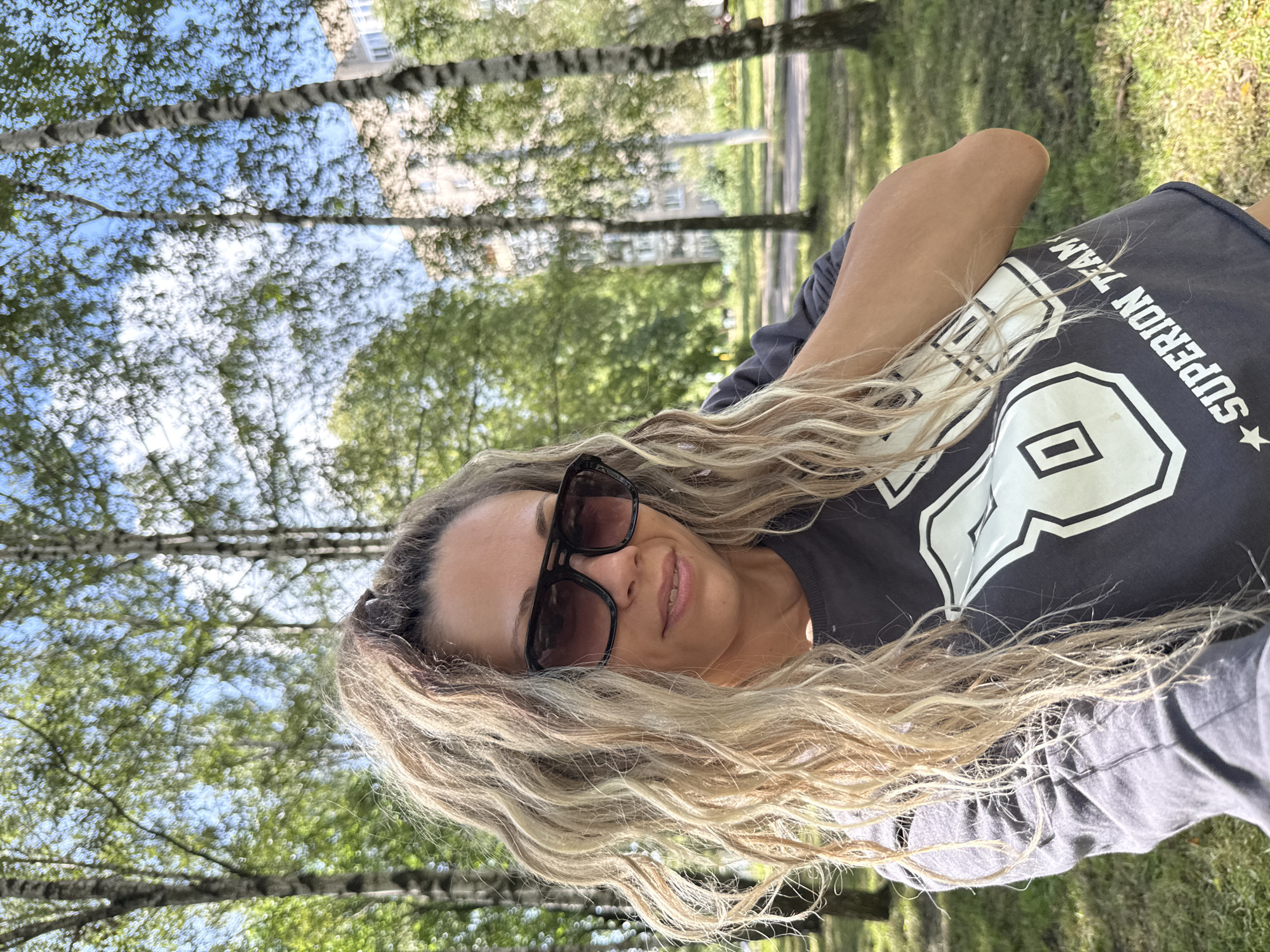 A woman with long, blonde, wavy hair, wearing sunglasses and a dark gray t-shirt with white lettering, is captured in a selfie. She is outdoors in what appears to be a park or residential area. Behind her, there are numerous birch trees with dappled sunlight filtering through their leaves. In the background, a multi-story building is visible, suggesting an urban or suburban setting. The ground is covered in green grass, with a paved pathway or road in the middle ground. The overall lighting indicates a bright, sunny day. The location is specified as Rusanovo, Russia.