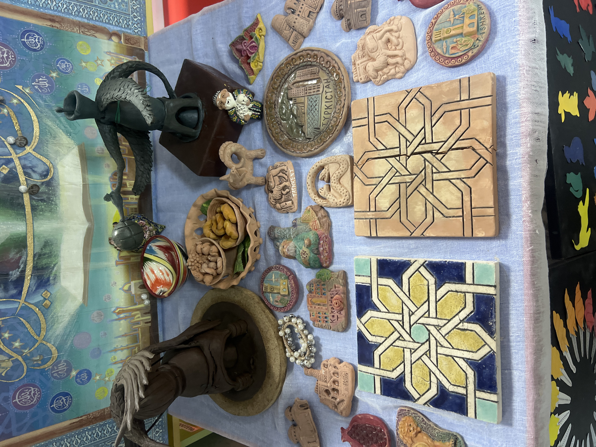 An overhead, slightly angled medium shot captures a display of artisanal souvenirs arranged on a blue cloth atop a table. The souvenirs are varied, including ceramic tiles featuring geometric patterns and depictions of architectural landmarks, small clay animal figures, a pomegranate-shaped ornament, and a bracelet. In the background, a vibrant tapestry with calligraphy and depictions of buildings hangs on a wall. The objects are diverse in color, texture, and material, suggesting they are handcrafted items from different regions.