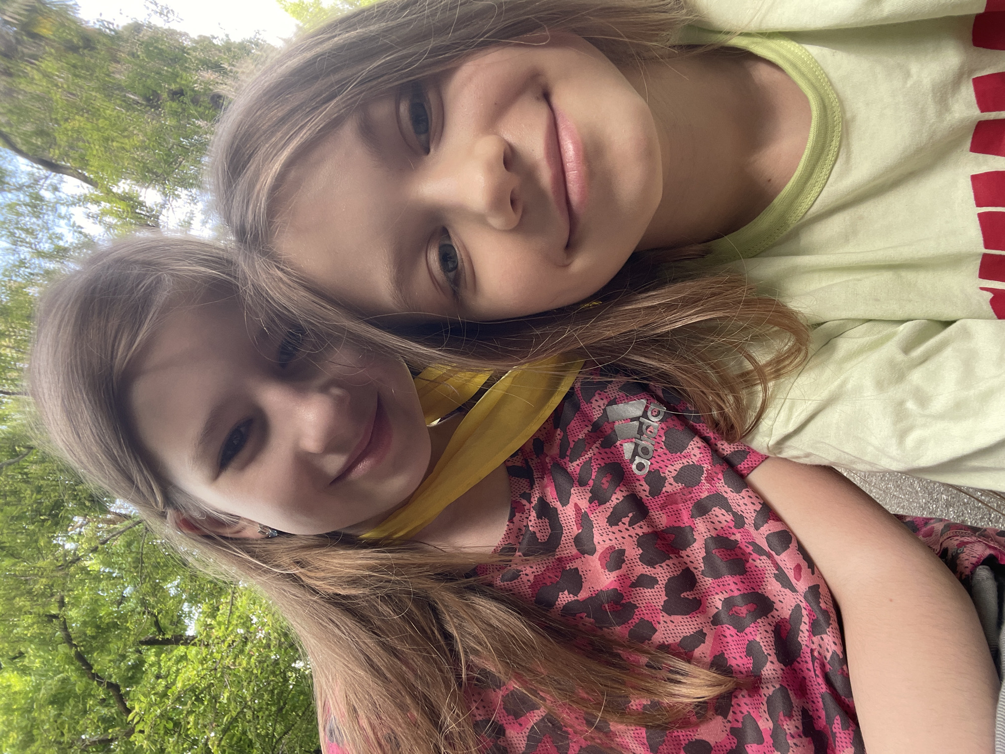 Two young girls are closely positioned together, likely in an outdoor setting. One girl is wearing a light green shirt with a light green neck trim and red text, while the other wears a pink and dark gray spotted shirt with the Adidas logo visible on the shoulder. A yellow object, possibly a scarf or piece of cloth, is draped around the girl in the spotted shirt. The background shows glimpses of foliage, suggesting a park or wooded area. The girls are smiling and appear to be taking a selfie or picture together. There is no evidence in the image to suggest it was taken in Sochi, Russia.