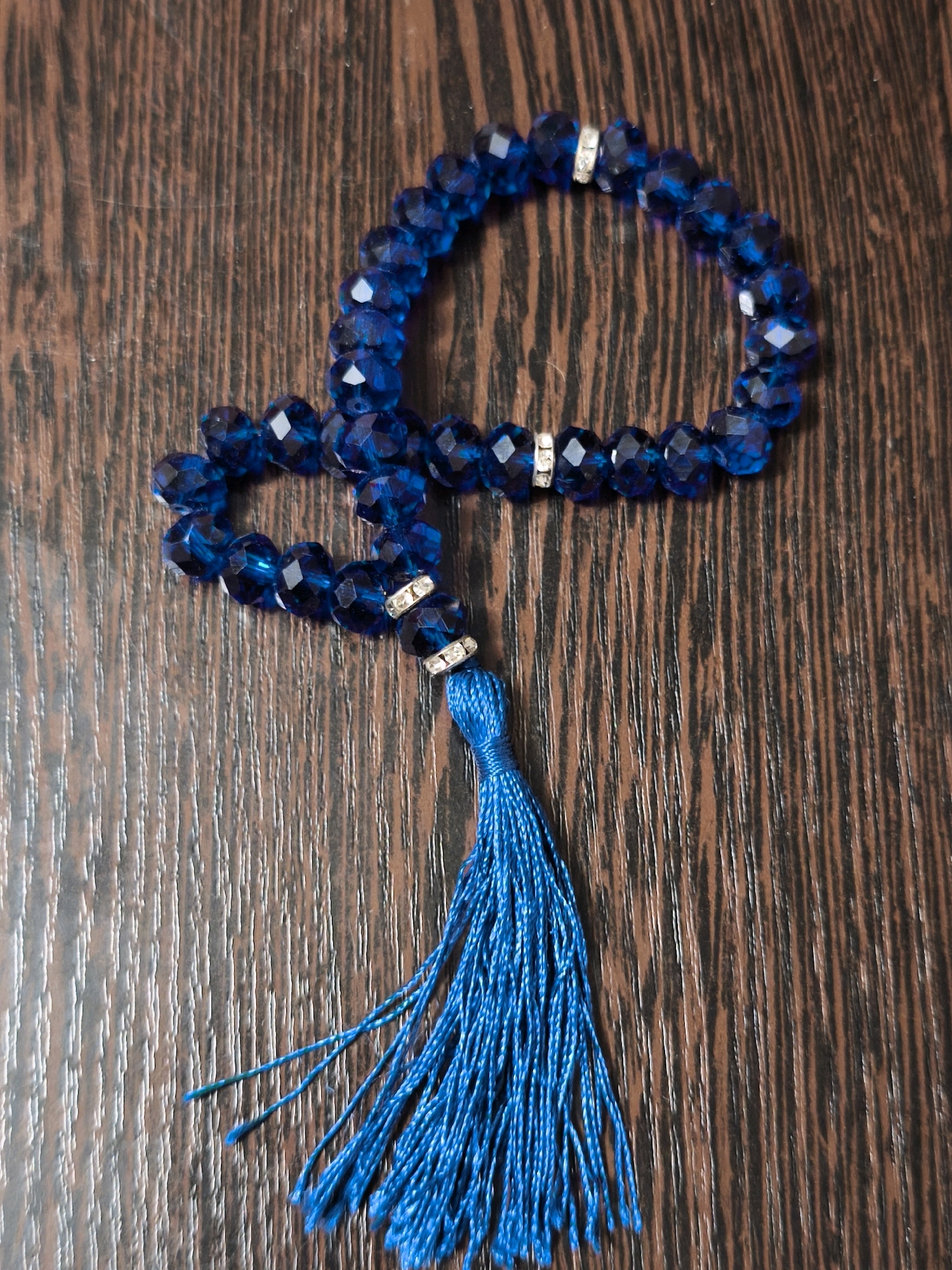 A close-up, overhead shot shows a strand of blue crystal beads with a tassel hanging from it, placed on a dark brown, wood-grained surface. The beads are faceted and appear to be connected by an elastic string, forming a loop that is partially visible. There are two small, decorative metal rings with clear, faceted stones interspersed between the beads. The tassel is also made of blue thread and hangs loosely. The wood grain on the surface is distinct, with vertical lines and variations in tone. The image is well-lit, with some shadows cast by the beads. There are no people or animals visible in the image, and no indications of the specific location in Karachayevsk, Russia.