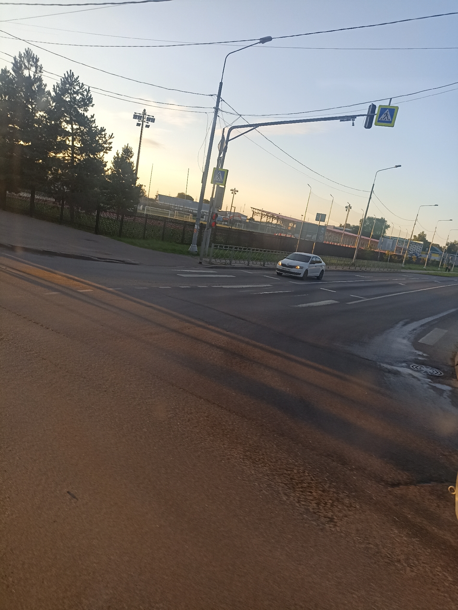 The image captures a street scene in Chekhov, Russia, likely during late afternoon or early evening, judging by the low angle of the sunlight casting long shadows. The foreground is dominated by a paved road with distinct lane markings. A white car is visible in the middle ground, driving on the road. The background features a pedestrian crossing, traffic signals, and overhead wires. Beyond that, there's a sports field with lighting towers and a dark-colored fence. Trees and other structures are also present in the distance. The sky is clear and light blue, suggesting fair weather.