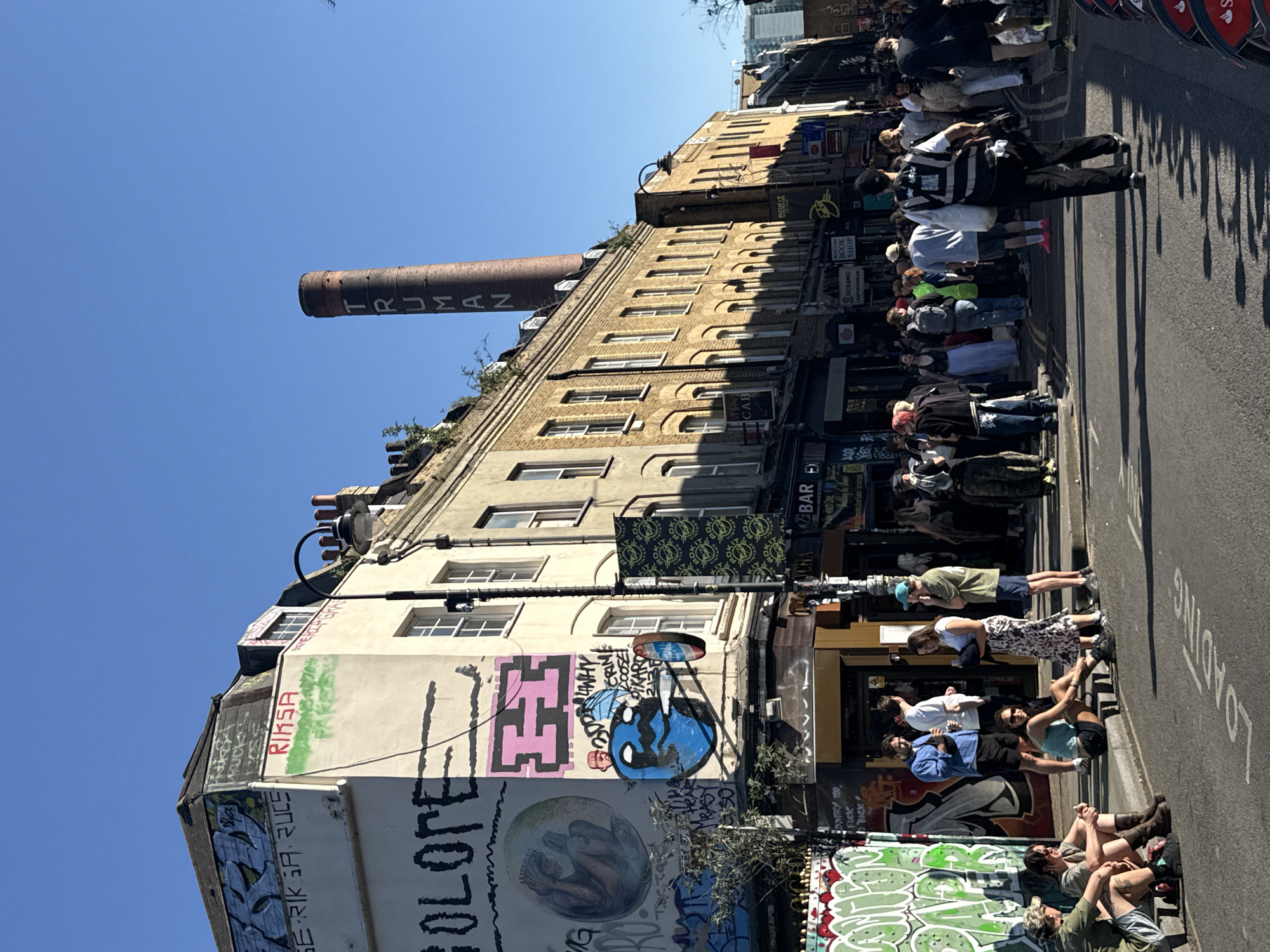 The image shows a street scene in London, United Kingdom. The street is lined with buildings, some with graffiti. A tall chimney with the name "TRUMAN" on it is visible. People are standing and sitting on the sidewalk and street. Some of the buildings have signs for bars and shops, including "OCEANIC BOOK SHOP" and "CAB". The street has the word "LOADING" painted on it. The sky is blue.