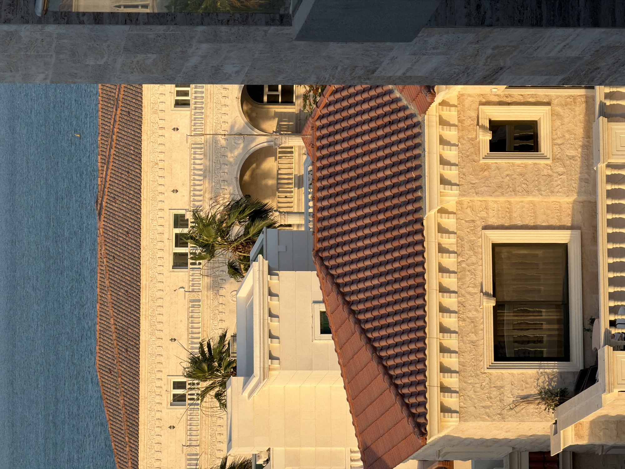 The image captures an elevated view of a coastal architectural scene, likely during sunrise or sunset, given the warm, golden light and prominent shadows. In the upper background, a vast, calm expanse of deep blue water stretches horizontally across the frame. A small, yellow-colored object, possibly a buoy, is faintly visible on the water surface in the upper-right quadrant.
In the midground, a large building with a reddish-brown, Spanish-style terracotta tiled roof dominates the central-left. Its facade is light beige or cream-colored, textured, and adorned with elaborate classical-inspired architectural details, including a prominent cornice with repetitive square motifs. Below the roofline, a continuous balustraded balcony is visible, behind which are several arched openings. Palm trees with green fronds are scattered between and around the buildings, illuminated by the sunlight.
In the lower foreground, another building with a similar reddish-brown terracotta tiled roof is positioned slightly lower and closer to the viewer. Its facade is also light-colored and textured, featuring decorative window frames. A large, rectangular window with dark, reflective glass is present on the left, reflecting a white balustrade. To its right, a smaller, square window is visible. A white balustrade, likely part of a terrace or another balcony in the immediate foreground, is partially visible at the bottom-right. To the left, a flat-roofed section of a building with clean lines and a distinct cornice is visible.
A prominent, vertical, light-colored stone column or wall element frames the right side of the image, partially obscuring the buildings behind it. This structure exhibits horizontal striations in its texture, and a dark, reflective surface (possibly a window) within a recessed area reflects green foliage.
The overall architectural style, combined with the presence of palm trees and a large body of water, suggests a coastal region with a Mediterranean, Middle Eastern, or similar warm climate. The exact location in the city of Unknown, Unknown cannot be determined from the visual information provided. No people are visible in the scene.