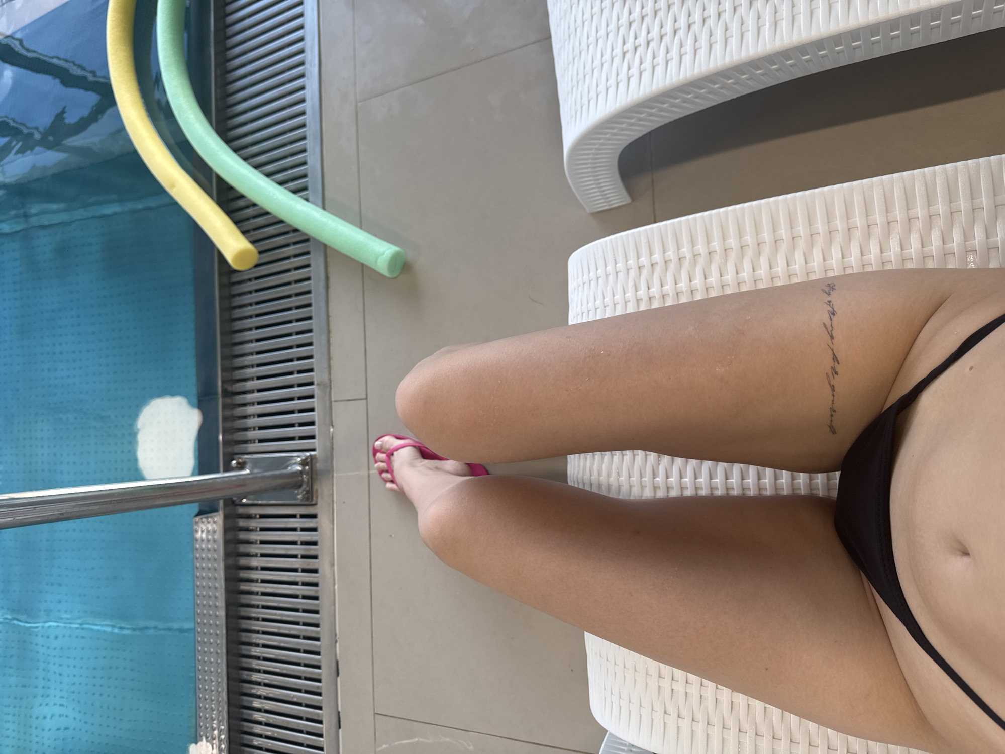 The image shows a person's legs and torso from a first-person perspective, lying on a white lounger next to a swimming pool. The person is wearing a black bikini and pink flip-flops. There are two pool noodles, one yellow and one green, near the edge of the pool. The pool is filled with blue water and has a metal railing. The ground around the pool is tiled. The image does not provide any information about the city of Chaikovsky, Russia.