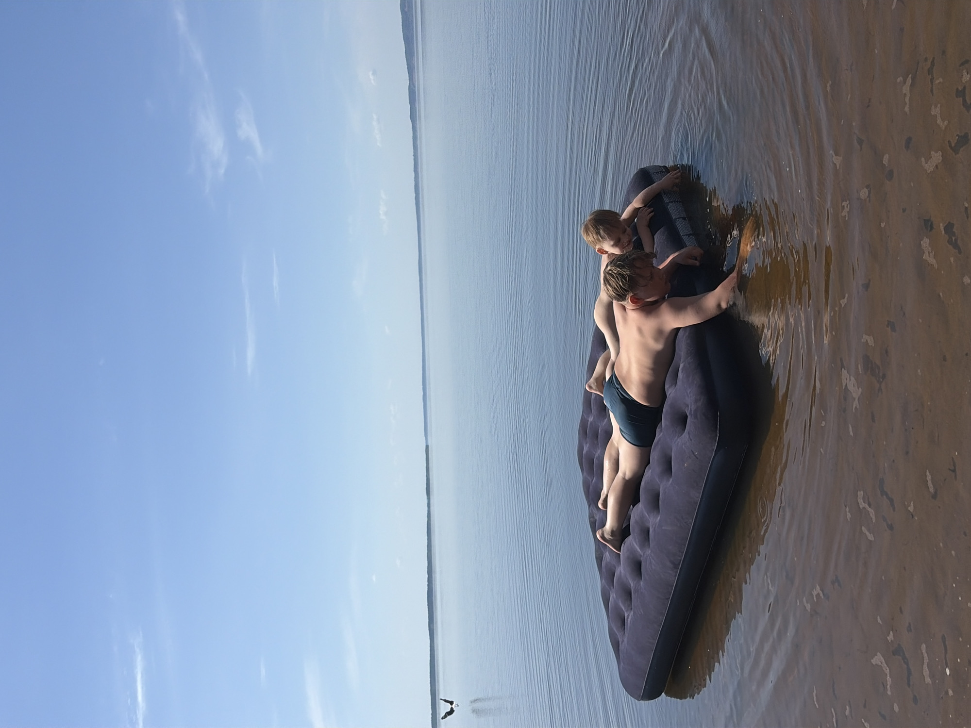 Two shirtless children are on an inflatable mattress in a body of water. The mattress is partially beached on a sandy area. The location is Zabegaevo, Russia, as inferred from the prompt. The sky is blue with scattered clouds, and the water extends to the horizon with a darker line of land visible in the distance.