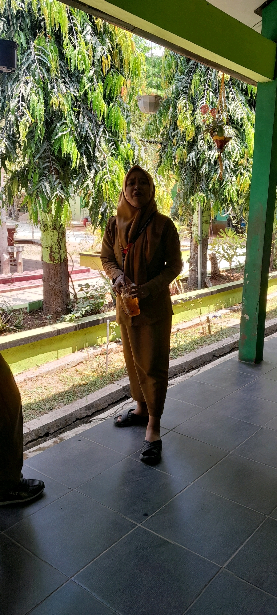 In Tahunan, Indonesia, a woman wearing a tan hijab and a tan uniform stands outside, holding a clear cup with a beverage. She is positioned in front of a building with a green painted wall and a white overhang, suggesting a school or government facility. Tall trees with lush green foliage surround the area, creating a natural backdrop. The ground is paved with dark tiles.