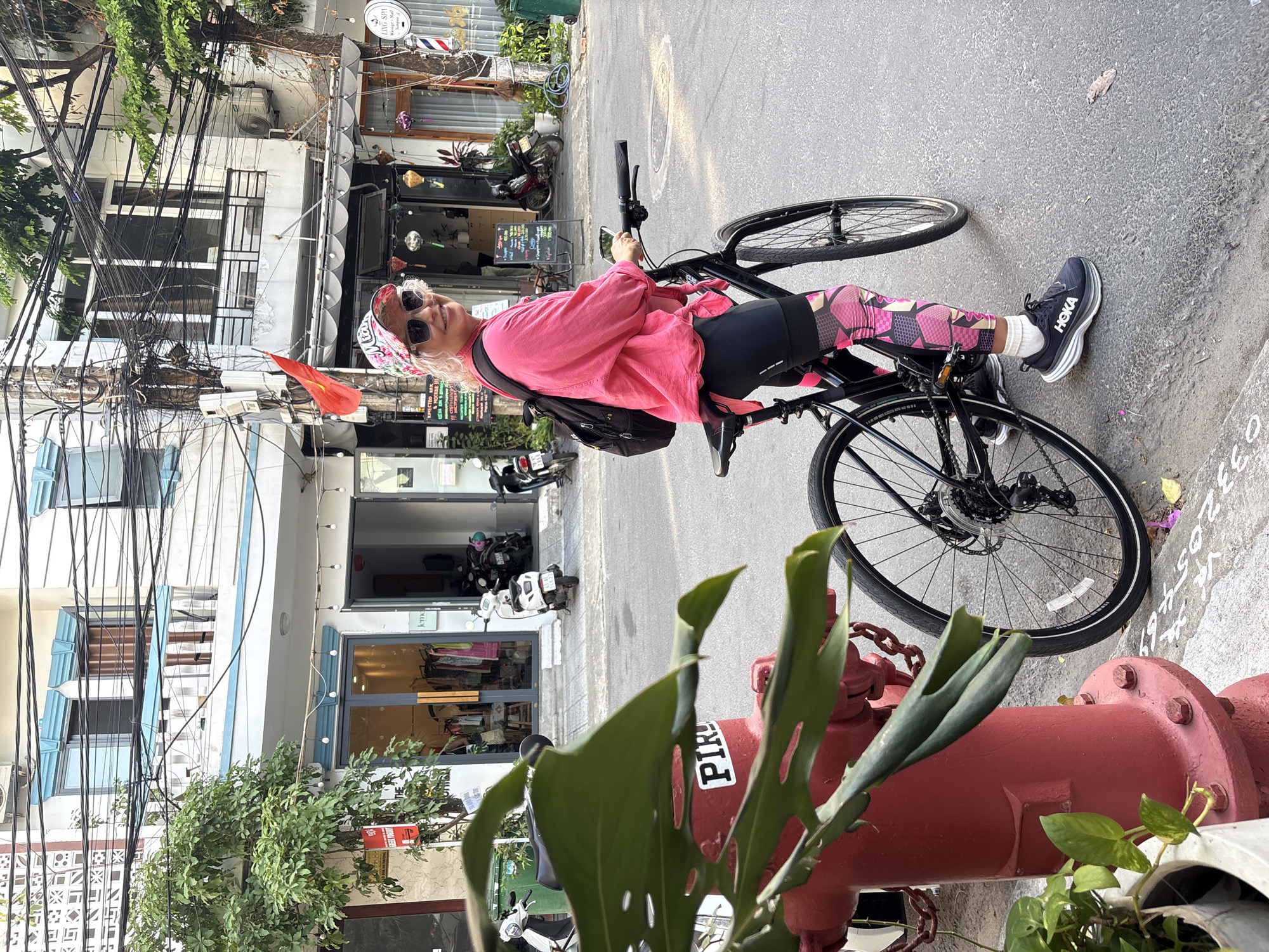 A woman sits astride a black bicycle on a sidewalk next to a red fire hydrant, in what appears to be a city street scene. She is wearing a pink shirt, patterned leggings, black sneakers marked with the word "HOKA" in white letters, sunglasses, and a floral patterned head covering. A black backpack is slung over her shoulder.
The background shows a variety of buildings with storefronts. Electrical wires run haphazardly across the sky above the street. Motorcycles are parked along the sidewalk. Vietnamese writing can be seen on the sidewalk and signage. The location appears to be Đà Nẵng, Vietnam.