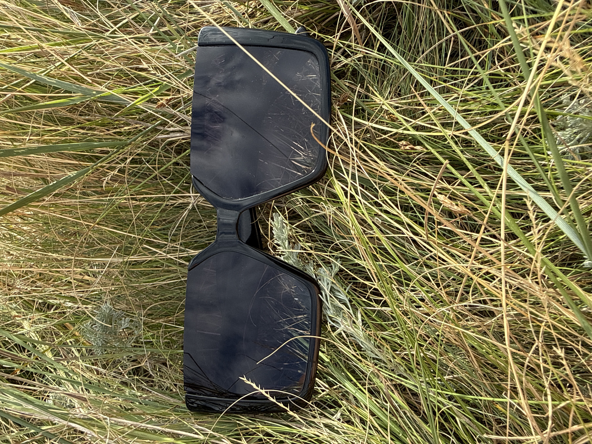 A pair of black, oversized sunglasses is lying on the ground amidst tall, dry grass. The sunglasses have a thick, angular frame and dark, reflective lenses. The lenses show scratches and reflections of the surrounding vegetation. The grass is a mix of green and golden-brown hues, appearing dry and wind-swept. A small, pale grey plant is visible among the grass near the left lens of the sunglasses. The scene suggests an outdoor environment, possibly a field or roadside in a rural area. The provided metadata indicates the location is in the city of Өндіріс, Kazakhstan.