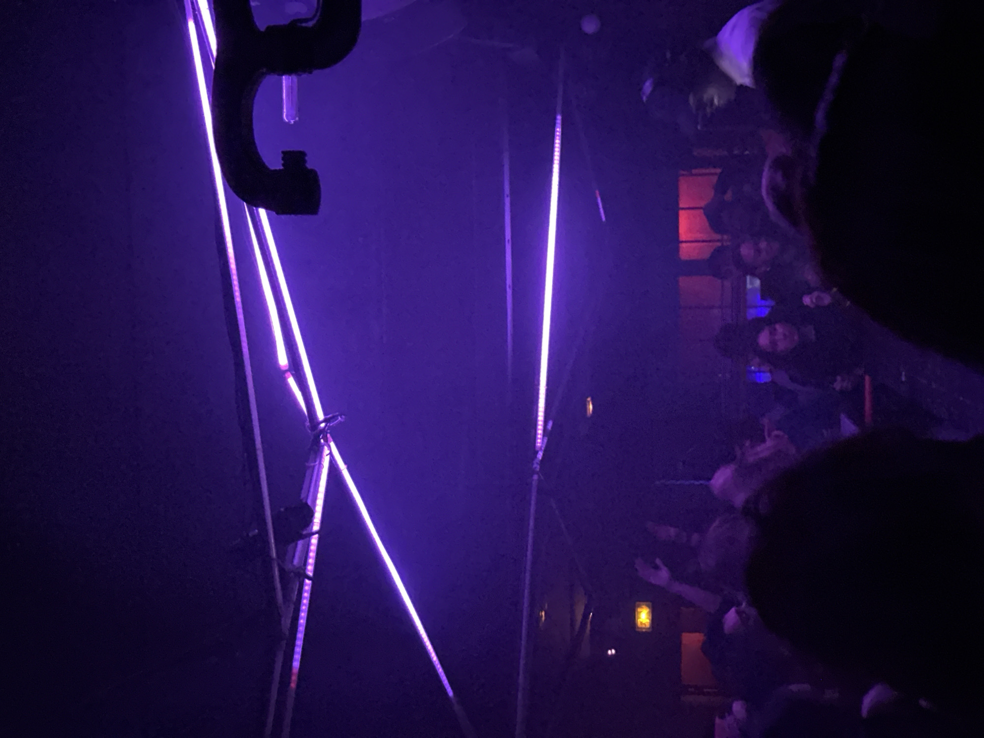 An indoor nightclub or music event is depicted in a dark, enclosed space, likely in Paris, France. The environment is illuminated predominantly by multiple purple LED strip lights affixed to the ceiling, some running diagonally. A dark, curved metal pipe, possibly part of ventilation or a lighting rig, is visible in the upper right quadrant. An emergency exit sign, illuminated in yellow, is present on a wall in the mid-left background. A noticeable atmospheric haze or smoke permeates the space.
A crowd of individuals occupies the lower and mid-ground, their forms largely silhouetted and indistinct due to the low light. In the mid-ground on the left, one person has an arm raised. On the right side of the frame, a person wearing headphones, likely a DJ or performer, is discernible in a lighter-colored top, facing towards the crowd. Several red-lit rectangular panels or windows are visible in the distant background. The specific address or name of the venue within Paris is not ascertainable from the image.