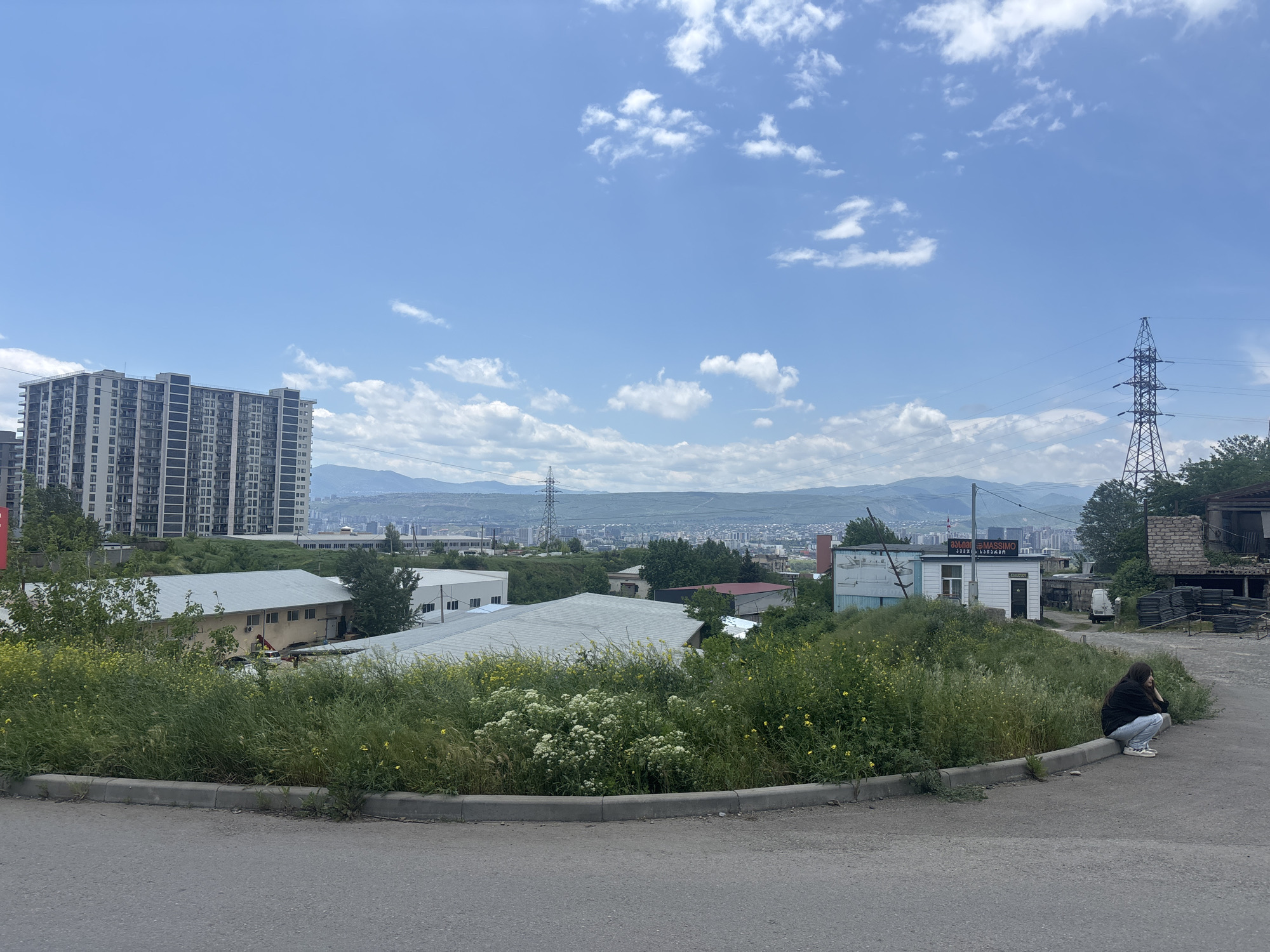 The scene is an urban landscape in Tbilisi, Georgia, on a sunny day with a partly cloudy sky. In the foreground, a person wearing dark clothing and light-colored pants sits on the edge of a low concrete barrier along a paved road. Behind the person is overgrown vegetation with yellow and white flowers.
The midground features a mix of industrial and residential buildings. To the left, there is a tall, modern apartment building and several one or two-story industrial buildings with light-colored roofs. To the right, a small white building with a sign in Georgian script is visible, along with remnants of a dilapidated brick structure, discarded tires, and a utility pole.
In the background, the cityscape of Tbilisi stretches out towards the distant mountains. Power lines run across the scene, connecting the utility pole to the cityscape. The overall impression is a view of a city in transition, with modern buildings juxtaposed against older, less maintained structures.