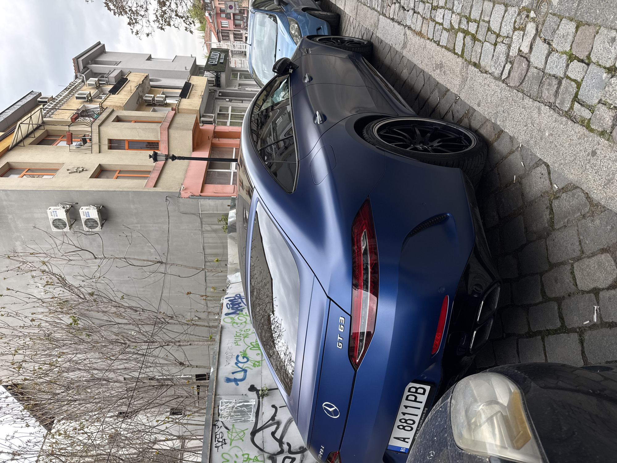This urban street scene, set in Plovdiv, Bulgaria, captures a row of parked cars alongside multi-story buildings under an overcast sky. The image appears to be taken during daylight hours in what seems to be late winter or early spring, given the bare tree branches just beginning to show nascent green leaves.
The main subject is a dark blue, matte-finish Mercedes-AMG GT 63 4-Door Coupe, parked facing away from the viewer on a cobblestone street. Its rear is prominently featured, displaying the "GT 63" badge and the Mercedes-Benz emblem. The Bulgarian license plate, "BG A 8811 PB," is clearly visible, along with the text "www.AutoHaus.bg". The car is fitted with distinctive black multi-spoke wheels. In the immediate foreground, part of a darker vehicle, possibly black, shows a dirty and aged headlight. Behind the Mercedes, a light blue hatchback is parked further down the street.
To the left, a series of multi-story buildings line the street. These structures vary in architectural style and color, with facades in shades of beige, grey, and some with reddish or pinkish accents. Many buildings feature multiple external air conditioning units and small balconies. A black lamppost stands vertically near the buildings.
The street and sidewalk are paved with worn cobblestones, with a defined curb separating the parking area. Scattered cigarette butts are visible on the cobblestones near the parked cars. A wall or fence behind the Mercedes is covered in various spray-painted graffiti. A commercial sign on one of the background buildings reads "GALARDO REAL ESTATE". The scene is static, depicting a typical urban environment.