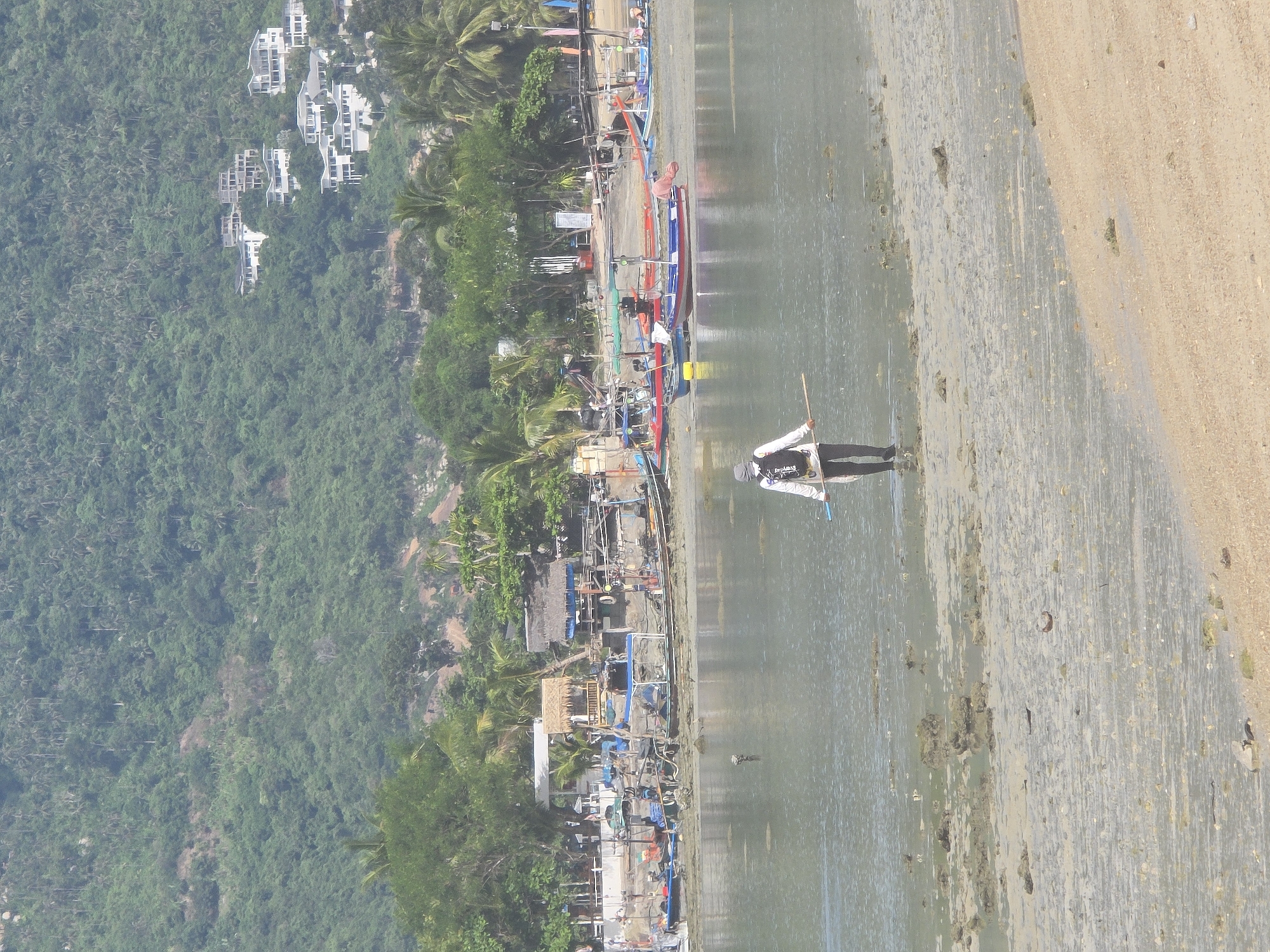 The image depicts a coastal scene in Baan Lamai, Thailand. In the foreground, a person stands in shallow water, holding a long pole. This individual is dressed in a white long-sleeved shirt, dark pants, and a backpack. The water is calm and reflects the sky, with the sandy seabed visible beneath the surface, scattered with rocks and debris.
In the middle ground, a line of colorful fishing boats is moored along the shore. These boats vary in size and color, with some displaying canopies or awnings. The shoreline itself is a mixture of sand and mud, with various structures and vegetation visible further back.
The background features a densely forested hillside, characteristic of tropical landscapes. Several modern white buildings are nestled into the hillside, suggesting a developed area within the natural environment. The overall impression is a tranquil day at a Thai beach, with evidence of both local fishing activities and tourist infrastructure.