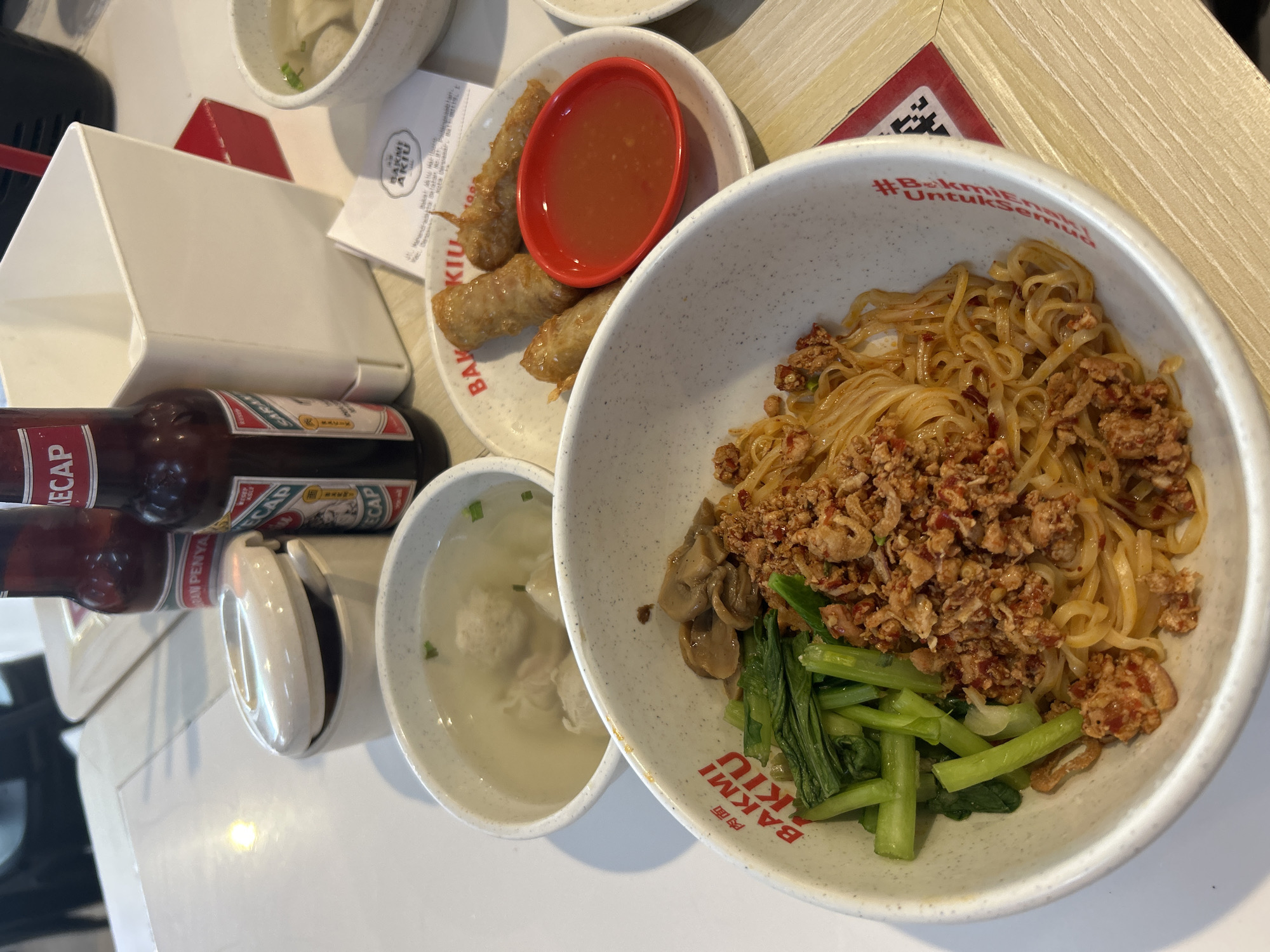The image shows a close-up of a table at Bakmi Akiu, a restaurant located in Denpasar, Indonesia. The focus is on a bowl of noodles topped with minced meat and vegetables. Next to it, there is a small plate with three fried spring rolls and a dish of red dipping sauce. In the background, there are two bottles of Kecap (Indonesian soy sauce), a white holder, and a bowl of soup with meatballs. A receipt from Bakmi Akiu is also visible on the table. The table surface appears to be made of light-colored wood.