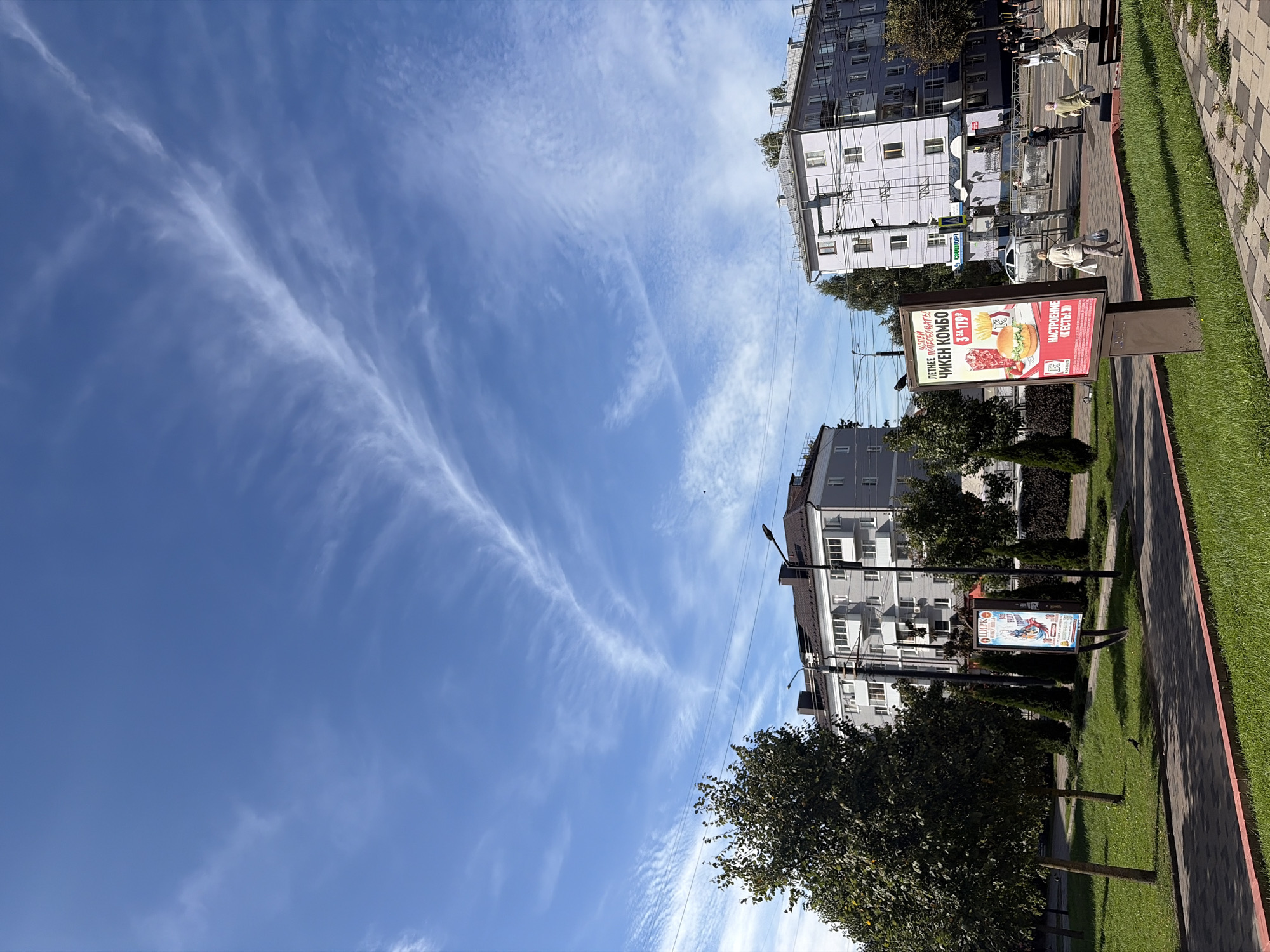 This image captures a daytime outdoor scene in Sof'ino, Russia. The sky is a vibrant blue, adorned with wispy cirrus clouds forming abstract patterns. In the foreground, a paved walkway with a red border borders a well-maintained grassy area. Several trees, some with full green foliage and others with slender trunks, are scattered across the lawn. A billboard advertising a "Chicken Combo" meal stands prominently on the right side of the walkway. Beyond the greenery, apartment buildings with light-colored facades and multiple stories line the street. A few people are visible walking along the sidewalk, and one person is on a bicycle. A streetlamp is positioned near the billboard. The overall impression is a snapshot of urban life on a clear day.