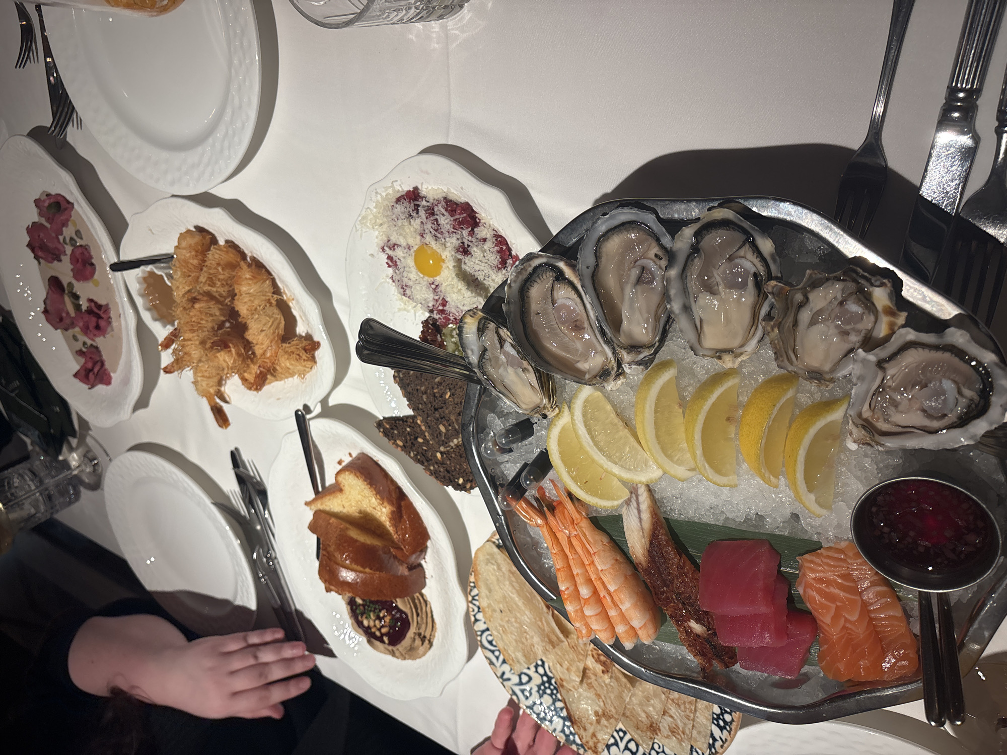 The image depicts an elaborate dining scene in an upscale restaurant, identified as being located in Saint Petersburg, Russia. The table is covered with a pristine white tablecloth and features a variety of meticulously prepared dishes, polished silverware, and glassware.
In the foreground, a large oval metal platter filled with crushed ice serves as a centerpiece, showcasing a chilled seafood and raw fish selection. This platter includes several fresh oysters on the half shell, a row of peeled cooked shrimp, slices of yellow lemon wedges, cubes of raw red tuna sashimi, slices of raw orange salmon sashimi, and a long piece of dark, cooked fish (possibly smoked eel) resting on a green leaf. A small metal cup containing a dark red sauce is also present on the platter. Small, dark, spherical objects (possibly caviar or balsamic pearls) are visible on the ice near the oysters.
To the upper left of the seafood platter, a white ceramic plate holds various flatbreads or crispbreads. Partially visible to the far left, a person's hand, with fair skin and wearing dark clothing, is reaching towards a different patterned plate. Another person's forearm and hand are visible in the upper left background, resting near an empty white plate.
Other dishes on the table include:
* On a white plate to the right of the bread, a mound of finely chopped red meat (beef tartare) is topped with grated white cheese and a raw egg yolk in its center. Several dark, seedy bread slices are positioned nearby.
* Above the beef tartare, a white plate contains several pieces of crispy fried shrimp, seemingly wrapped in phyllo or kataifi pastry, accompanied by a small container of dipping sauce.
* Further in the background, another white plate holds slices of a golden-brown bread (possibly brioche or challah) next to a dark spread garnished with pine nuts and what appears to be a red fruit compote or jam.
* In the upper background, a white plate features pink or red food items arranged in a creamy white sauce with small green garnishes, possibly a carpaccio or ravioli dish.
Multiple empty white ceramic plates, forks, knives, and various glasses (including water glasses and wine glasses) are distributed across the table, suggesting a meal in progress or recently served. The lighting appears to be indoor artificial light, casting some shadows.