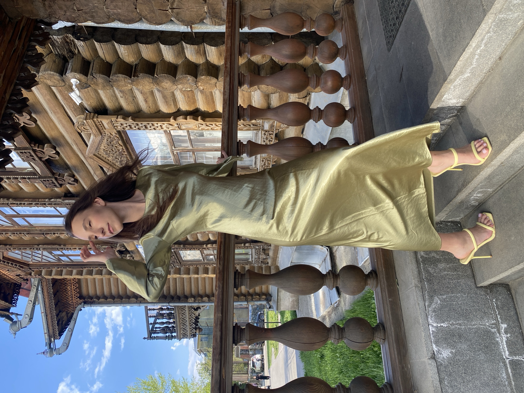 A slender woman in a floor-length, light-green gown and yellow sandals poses on a stone step in front of a railing. The ornate wooden architecture of a building stands behind the railing. The blue sky with white clouds are visible beyond the eaves of the building. The woman's dark hair is long, and she is looking towards the camera with her left hand raised in front of her face. A road, sidewalk, and green grass are visible beyond the railing. This location is in Yekaterinburg, Russia.