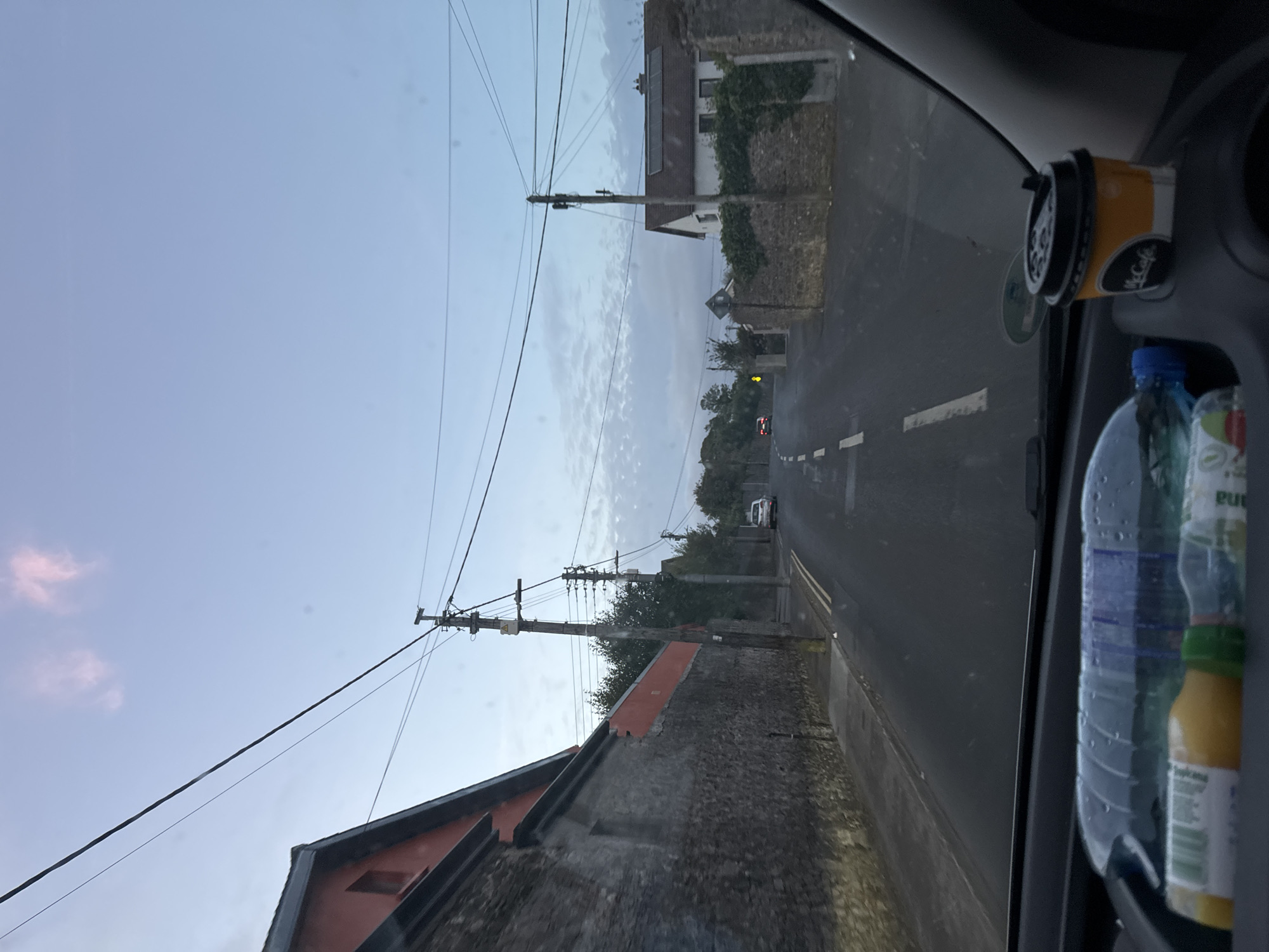 The image is taken from the interior of a vehicle looking out onto a street in Tramore, Ireland. The sky is a pale blue with some wispy pink clouds. There are overhead power lines visible across the sky, supported by wooden poles.
On the left side of the road, there is a long, low building with a red-tiled roof and a stone wall along its base. On the right side of the road, there is a more modern-looking white house with a dark roof, partially obscured by vegetation. The road itself is asphalt and appears wet, with white dashed lines marking the lanes. A few cars are visible in the distance, and a white van is parked on the side of the road.
In the foreground, inside the vehicle, a plastic water bottle and a bottle of orange juice are visible on the dashboard. A McCafe coffee cup with a black lid and straw is also on the dashboard. The overall impression is that of a slightly overcast day, likely early morning or late afternoon, in a residential or semi-rural area of Tramore.