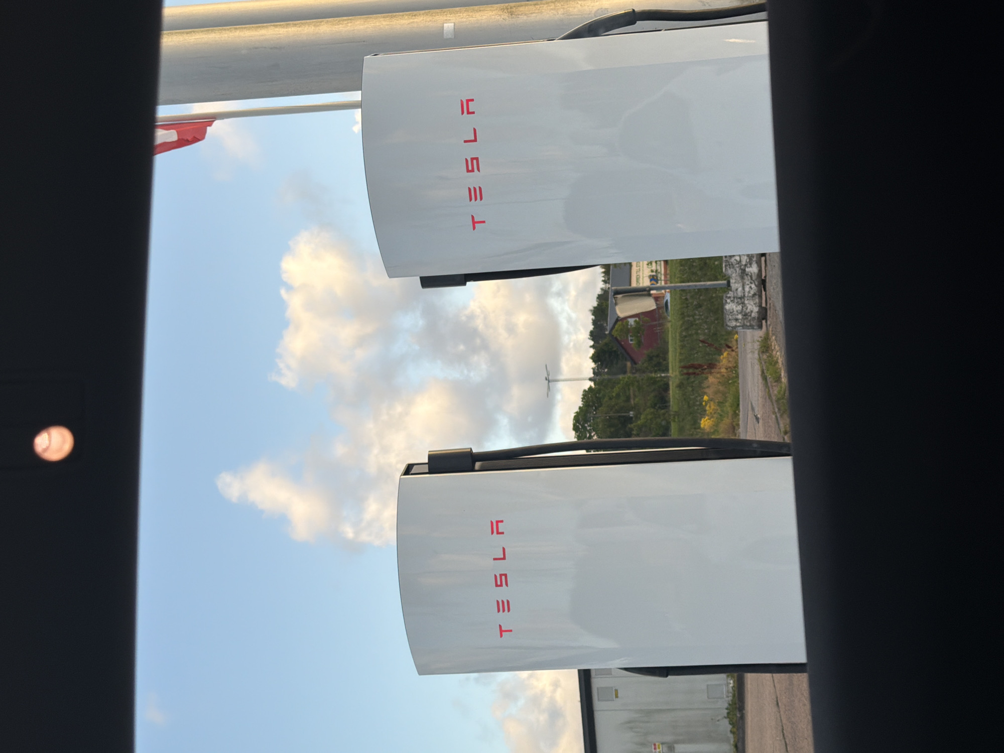 The image shows a Tesla Supercharger station in Hogstorp, Sweden, under a cloudy sky. Two white Tesla charging stations are visible in the foreground, each with the red Tesla logo. In the background, there is a building with a red roof, some trees, and a flagpole with a red and white flag. The ground is paved with asphalt and there are some yellow flowers. The image appears to be taken from inside a vehicle, looking out through the windshield. A blurry baseball or similar object is visible in the upper center of the image, possibly a reflection or something on the windshield.