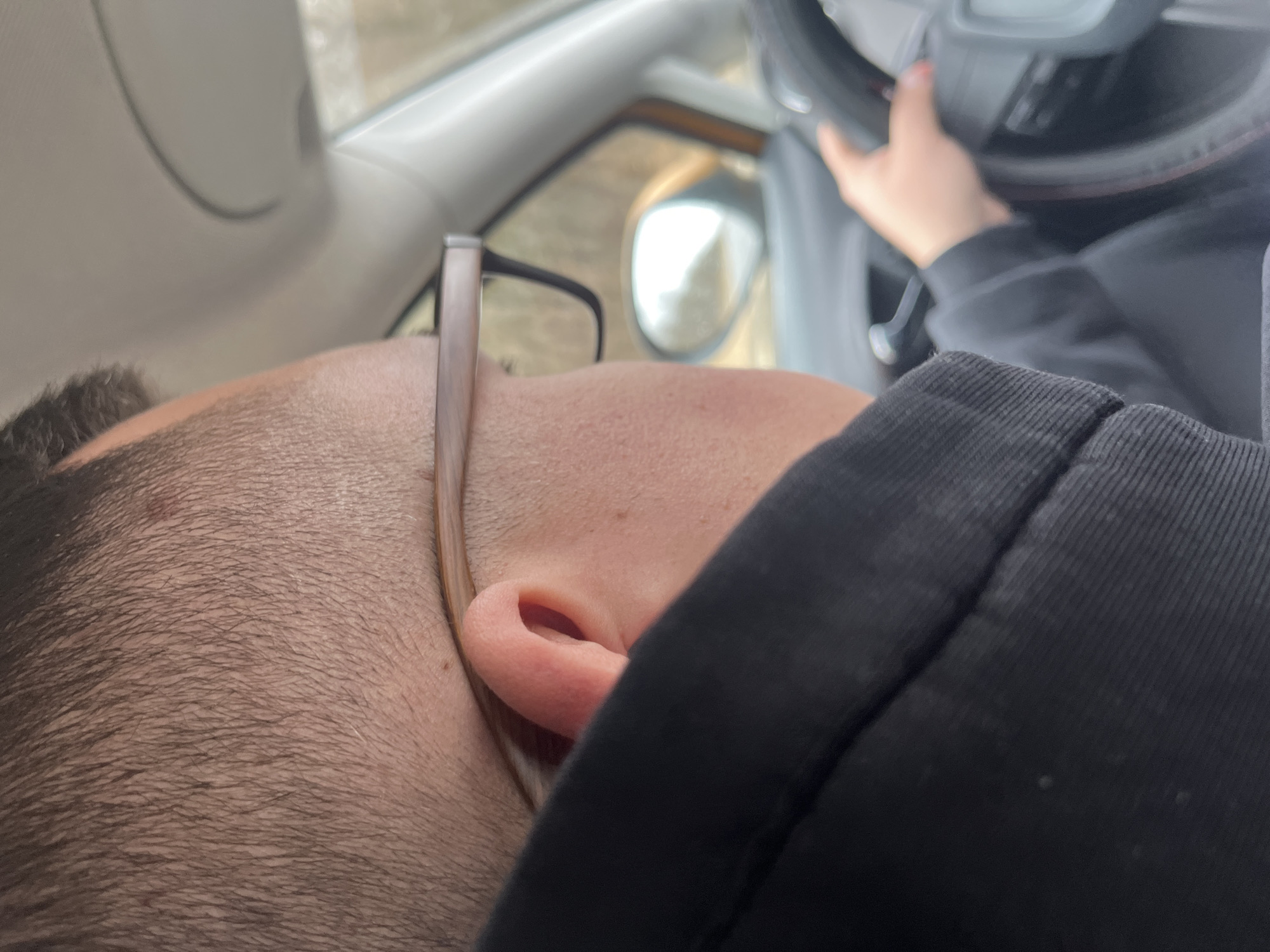 A man with dark hair and glasses is lying down inside a car. His head is near the driver's side window. A hand is visible on the steering wheel. The car's side mirror is visible in the background through the window. The interior of the car is light gray. The man is wearing a black hooded jacket. The location of Rogachovo, Russia cannot be determined from the provided image.