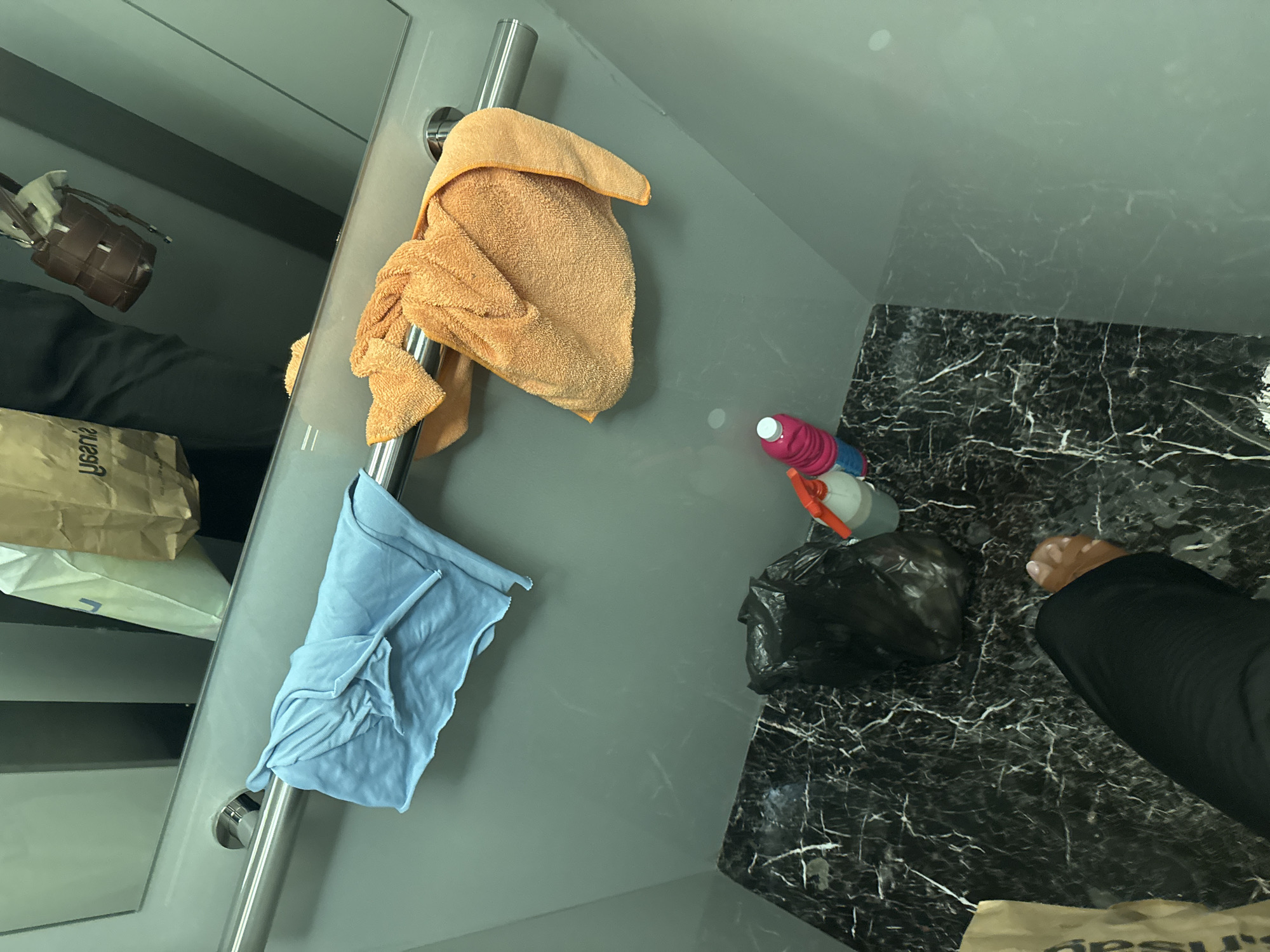 This image captures an indoor scene, likely a bathroom or utility space, in Burgas, Bulgaria, viewed from a high angle looking downwards. The flooring is a dark, black marble with prominent white veins. To the left, there's a smooth, grey wall or partition.
A chrome metal bar, possibly a towel rail or grab bar, is affixed horizontally to the grey wall. An orange, fluffy towel is draped over the top part of this bar, while a light blue cloth or towel is folded and hangs from the lower part.
On the marbled floor, a pink and white spray bottle, characteristic of cleaning products, is visible. Adjacent to it is a crumpled black plastic bag, which appears to be a trash bag. In the bottom right corner, a human foot with light-colored painted toenails is visible, belonging to a person wearing black trousers or leggings, suggesting they are standing in the space.
The reflective surface of the grey wall on the far left shows a blurred reflection of a person wearing dark clothing, holding a brown, woven bucket-style bag. Several paper bags are also visible in this reflection; one light brown bag has the partial text "ysania" on it, and another bag in the bottom right of the reflection shows the partial text "gesus".
The scene suggests a personal space, possibly a bathroom, with items related to cleaning or personal hygiene present. The time of day is not ascertainable, but the area is well-lit. There are no overt emotional cues.