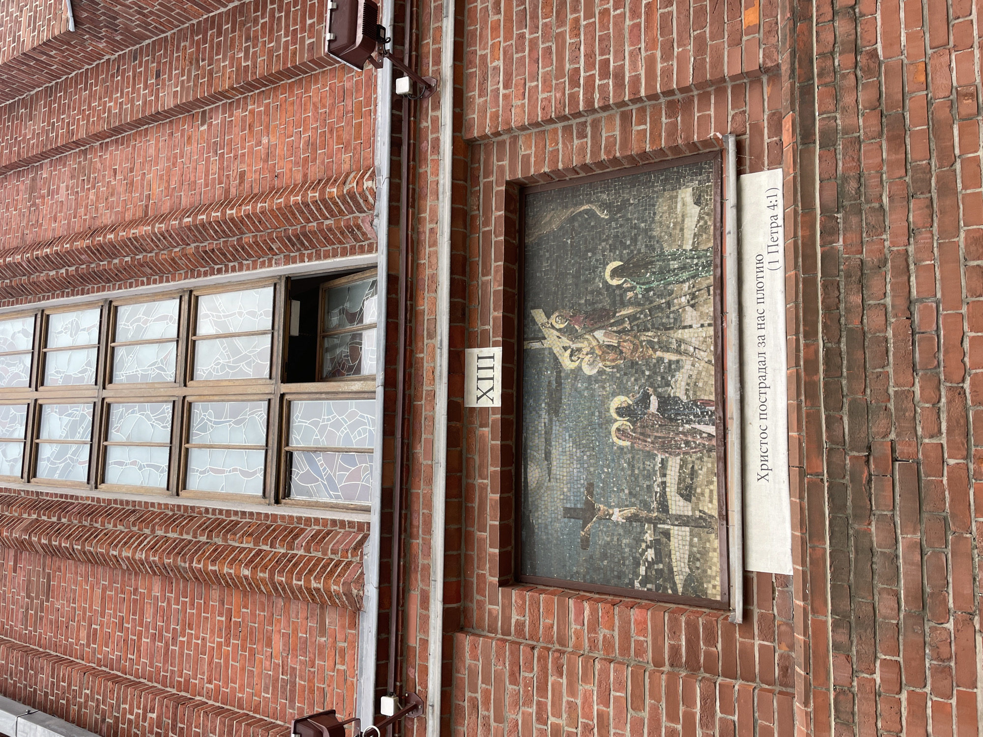 The image shows a close-up of a brick wall on the exterior of a building, likely a church given the mosaic artwork and stained glass windows. The wall is constructed of red bricks laid in a stretcher bond pattern, with decorative brickwork above a row of windows. The windows have wooden frames and feature stained glass with a white, abstract design.
To the right of the windows, a framed mosaic is mounted on the brick wall. The mosaic depicts a religious scene, likely the crucifixion of Jesus Christ, with figures in traditional religious attire. Below the mosaic, there is a white sign with dark text. A Roman numeral "XIII" is visible on a separate vertical sign to the left of the mosaic.
The visible text on the sign reads: "Христос пострадал за нас плотию (1 Петра 4:1)". This translates to "Christ suffered for us in the flesh (1 Peter 4:1)". The scene appears to be outdoors, with no indication of specific time of day or weather conditions. The overall impression is one of historical and religious significance.