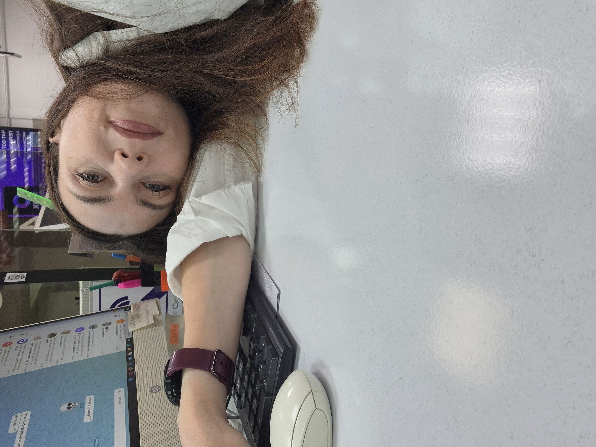 The media file shows a close-up, sideways view of a woman positioned at a desk in what appears to be an office or workspace. The image is rotated 90 degrees clockwise from its natural orientation.
**Subjects:** The central subject is a woman with long, brown hair and light skin, wearing a white collared shirt. Her face is clearly visible, with brown eyes, a subtle smile, and light pink lipstick. She wears a dark red or burgundy smart band on her left wrist. On the desk, there is a computer monitor displaying what looks like a messaging application or social media feed. An "HP" logo is visible on the monitor's stand. A dark keyboard and a white computer mouse are situated next to the monitor. A pen holder with colorful pens (blue, pink, green) is also visible behind the monitor.
**Scene and Setting:** The setting is an indoor professional environment, likely an office or study area, characterized by the desk, computer equipment, and office supplies. The desk surface is light-colored and slightly textured. The background behind the woman is somewhat blurred, showing indistinct office furniture or partitions, with a purple-lit sign.
**Activity:** The woman appears to be taking a casual selfie, looking directly at the camera, while at her desk. She seems to be pausing momentarily from an activity involving the computer.
**Notable Details:** The prominent feature is the image's orientation, presenting the woman and her surroundings as if viewed from a tilted perspective. The lighting is bright, creating a slight glare on the desk surface. On the computer screen, various profile pictures and chat bubbles are visible, with a distinct skull emoji prominently displayed in one of the messages.
**Visible Text:** The "HP" logo is clearly visible on the monitor stand. On the screen, chat messages are largely unreadable, but a skull emoji is evident. In the blurred background, inverted text that appears to read "TOP-TAP" and some Cyrillic characters are faintly discernible on a purple-lit sign.