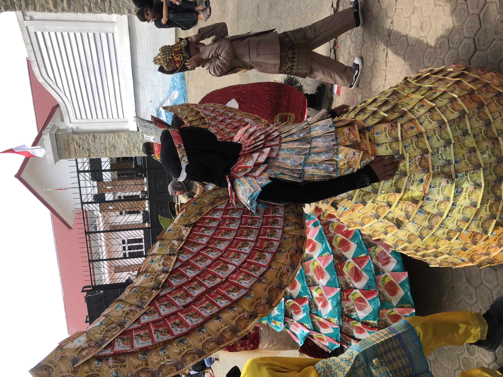 In Lhokseumawe, Indonesia, a procession takes place outdoors near a building with a red-tiled roof and stone accents, flying a red and white flag. A large, elaborate costume, possibly a parade float or wearable art, dominates the foreground. This costume is constructed from numerous small, rectangular packets, predominantly yellow with brown branding and red accents, and some teal packets featuring images of tuna. The costume has a layered, wing-like structure extending upwards and a voluminous, conical base. A person wearing a black head covering and long sleeves is partially visible behind the costume. To the right, a young boy in a formal brown outfit, complete with a decorative crown-like headpiece, walks past, touching his head. Other individuals are seated or standing in the background, some of whom appear to be spectators or participants. The ground is paved with interlocking grey stones. The overall scene suggests a cultural event or festival.