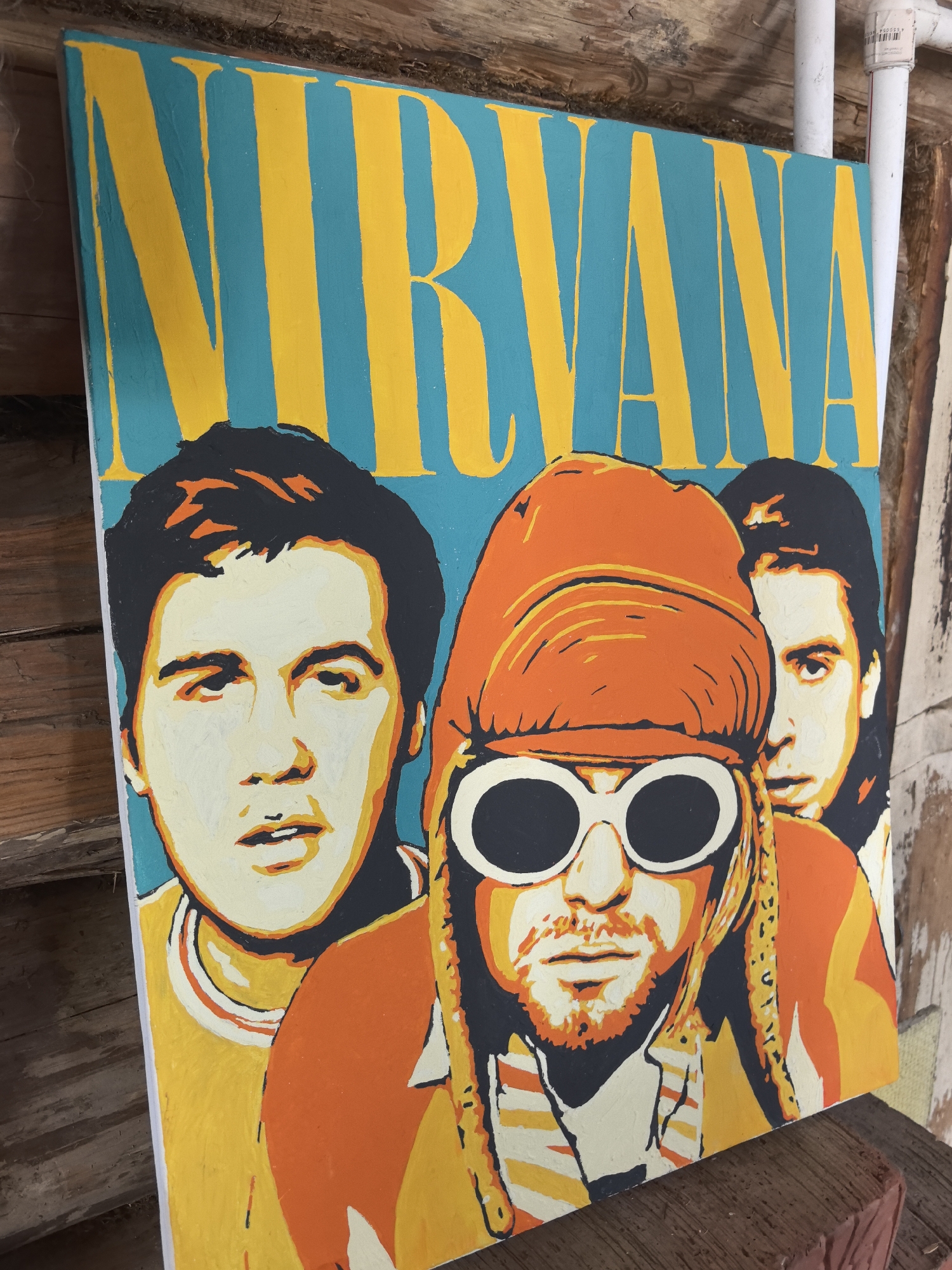 The image shows a painting of the band Nirvana. The painting is on a canvas with the band's name written in yellow letters against a teal background. Below the name are caricatured portraits of the band members, painted in shades of yellow, orange, black, and off-white. The painting appears to be resting on a wooden surface, possibly a shelf or ledge. Behind the painting is a wooden wall or structure, and a white PVC pipe is visible in the background as well. The location is not evident from the image.
