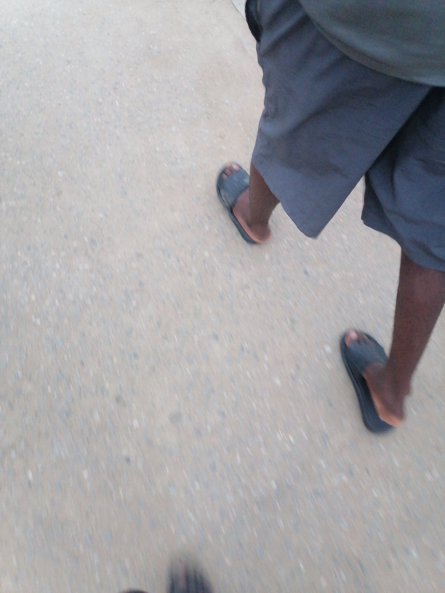 The image shows a close-up, low-angle view of two people's legs and feet walking on a paved surface. Both individuals are wearing dark-colored shorts and open-toed sandals. The ground appears to be asphalt or concrete with small stones embedded in it, consistent with road surfaces in Ikorodu, Nigeria. The lighting suggests it is daytime, possibly overcast, given the muted tones and lack of harsh shadows. The motion blur indicates the subjects are in motion. The scene captures a mundane moment of everyday life, likely on a street or pathway within the city.