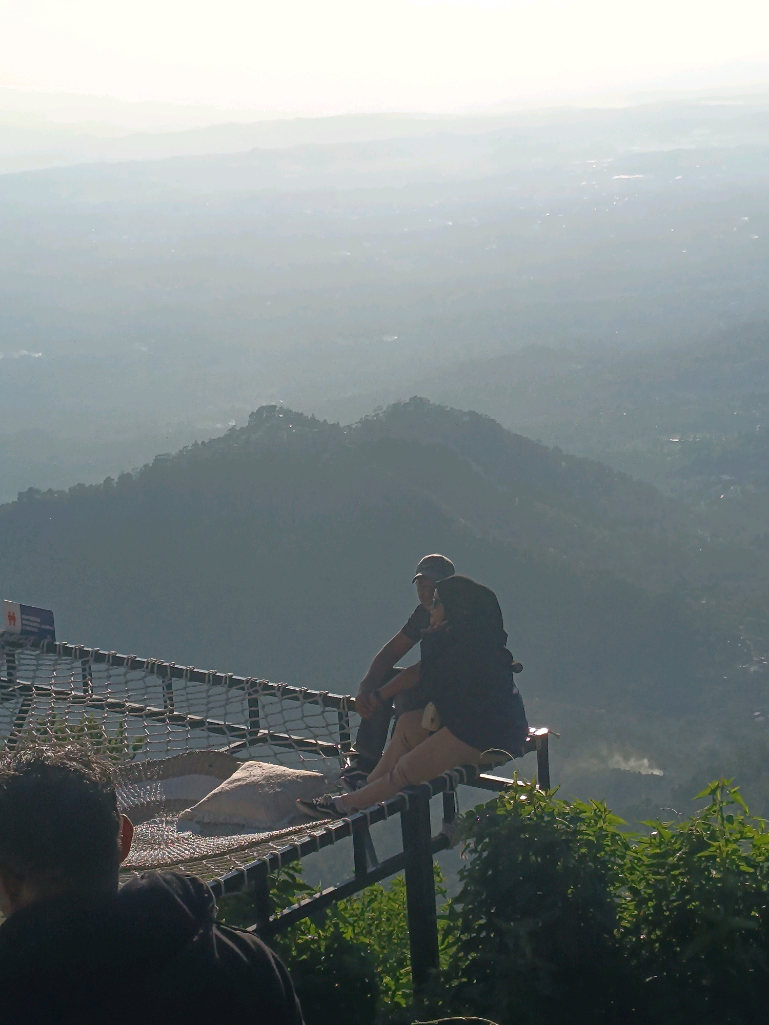 In the city of Keditan, Indonesia, two individuals are seated on a netted platform overlooking a vast, hazy landscape of rolling hills and distant mountains. The man, wearing a baseball cap and a dark t-shirt, sits with his arm around the woman, who is wearing a headscarf and a dark jacket, with her arm resting on his. They are both looking out at the expansive view. In the foreground, a portion of a person's head and shoulders, wearing a black hoodie, is visible from behind, also facing the view. The netted platform appears to be a lookout point, with a light-colored, irregularly shaped object, possibly a cushion or rock, resting on the net. Green foliage is visible at the bottom right of the frame, framing the scene. The sky is bright and overcast, contributing to the hazy atmosphere.