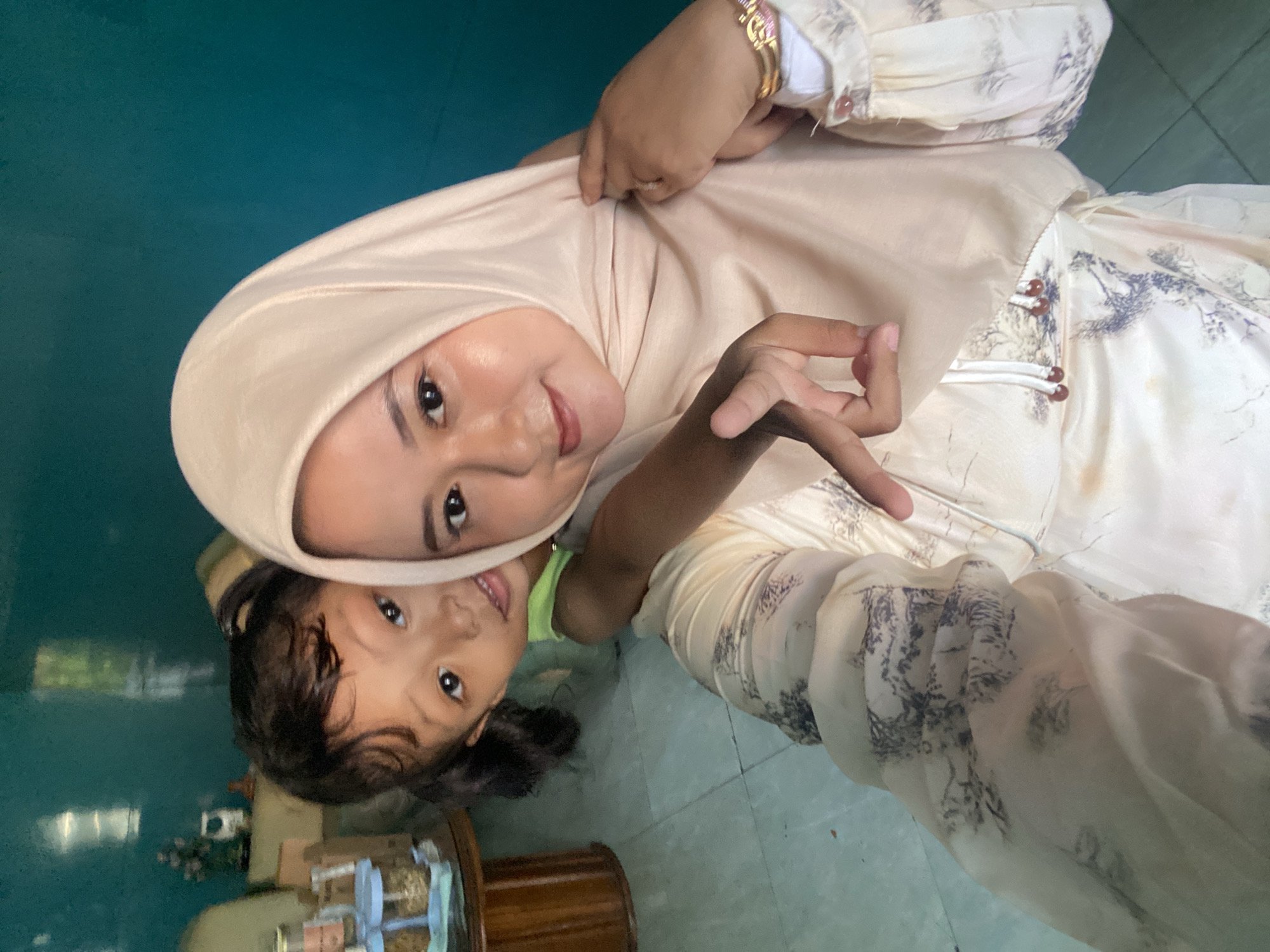 A woman wearing a light beige hijab and a dress with a light beige base and a pattern of gray-blue branches and leaves poses for a selfie with a young child. The woman is holding the edge of her hijab with one hand. The child, who is wearing a bright green shirt, is nestled near the woman. The background features a green-blue wall and a teal-colored floor. A dark brown wooden table with jars of various items sits in the lower left corner of the frame. The photograph appears to have been taken indoors, and the lighting is somewhat dim. The location is Gelam, Indonesia.