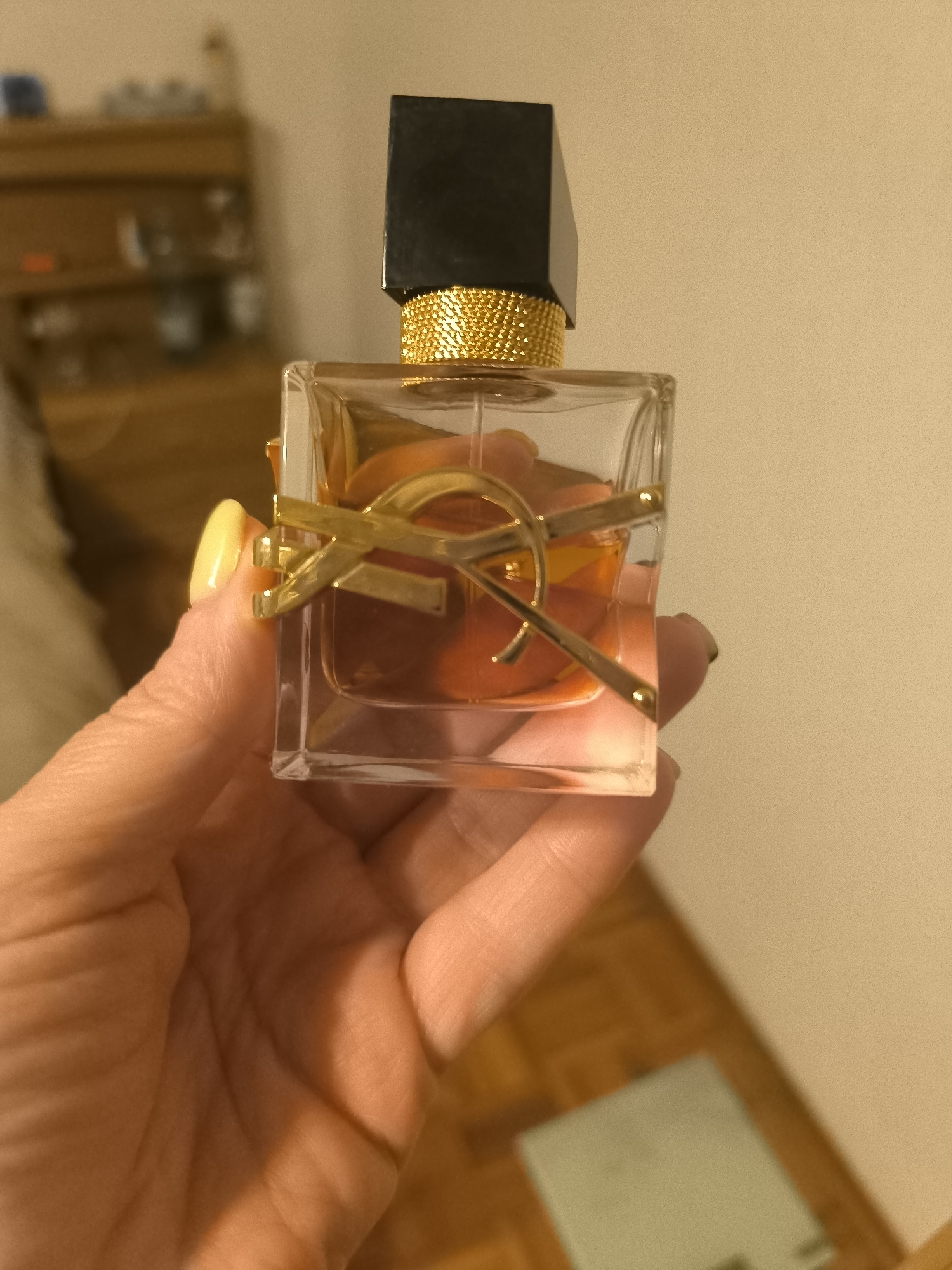 A hand with yellow nail polish holds a clear glass perfume bottle with a black cap and a gold Yves Saint Laurent logo. The perfume inside is an orange-pink color. The background is blurred, showing a wooden dresser and a light-colored wall. The scene appears to be indoors, possibly a bedroom.