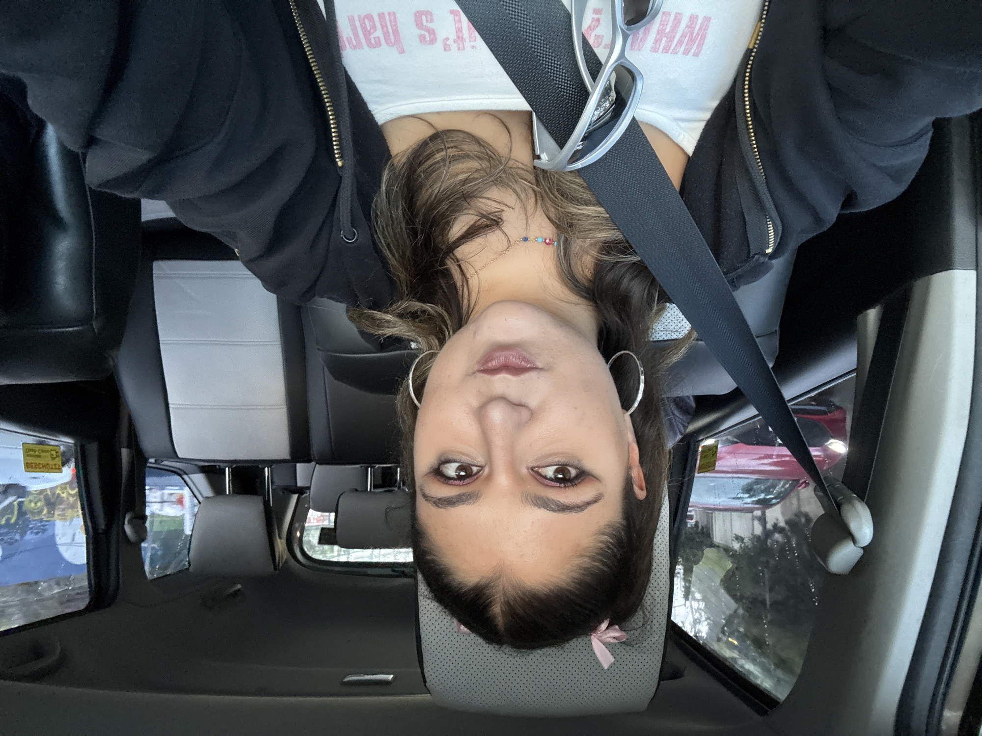 A woman is photographed from an upside-down perspective inside a car. She wears a white tank top with pink writing, a black zip-up hoodie, and large silver hoop earrings. A black seatbelt crosses her chest. Her brown hair is styled with a pink ribbon at the top of her head, resting against the car's gray headrest.
Outside the car window, a red vehicle is visible, along with foliage. Based on window stickers and the architecture observed outside the window, the location is identified as Mexico City, Mexico. The photo appears to have been taken from the driver's seat.
