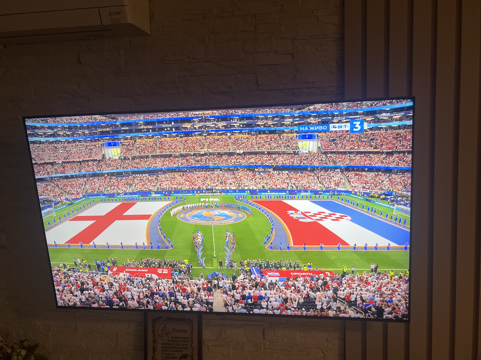 This image shows a television screen displaying a soccer match, likely a World Cup game, given the "FIFA WORLD CUP 2022" text visible on the screen. The screen is mounted on a wall covered with textured, light-colored stone or brick. To the right of the TV, there are vertical wooden slats.
On the screen, a large soccer field is visible, with a massive England flag and a Croatia flag laid out on the grass. A stadium filled with spectators wearing red and white clothing is also visible. A scoreboard at the top right of the screen indicates "НА ЖИВО" (LIVE in Russian) and shows the score as 3.
Below the television, a framed picture with text in Cyrillic script is mounted on the wall, suggesting the location is in a predominantly Slavic-speaking region. The text, which appears to be a religious passage or quote, is partially obscured but contains the word "ГОСПОДИ" (LORD in Russian). Given the presence of Cyrillic script, and the request for Varna, Bulgaria as a potential location, it is highly probable that this scene is taking place in a home or public space in Varna, Bulgaria. The overall ambiance is that of a viewing party or a domestic setting where a sporting event is being watched.