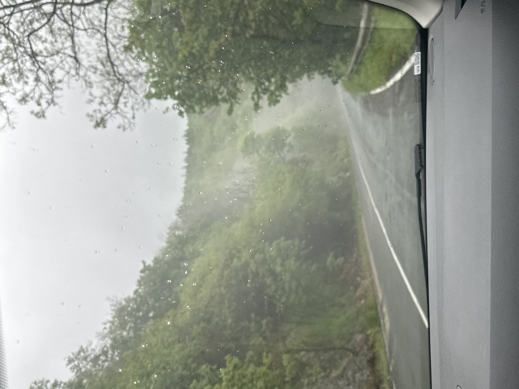 The image is taken from inside a vehicle, looking out through a windshield. Raindrops are visible on the glass. The view is of a road winding through a hilly, wooded area. The vegetation appears lush and green. A white line marks the edge of the road. The interior of the vehicle is visible on the right side of the image. The location is Karnare, Bulgaria.