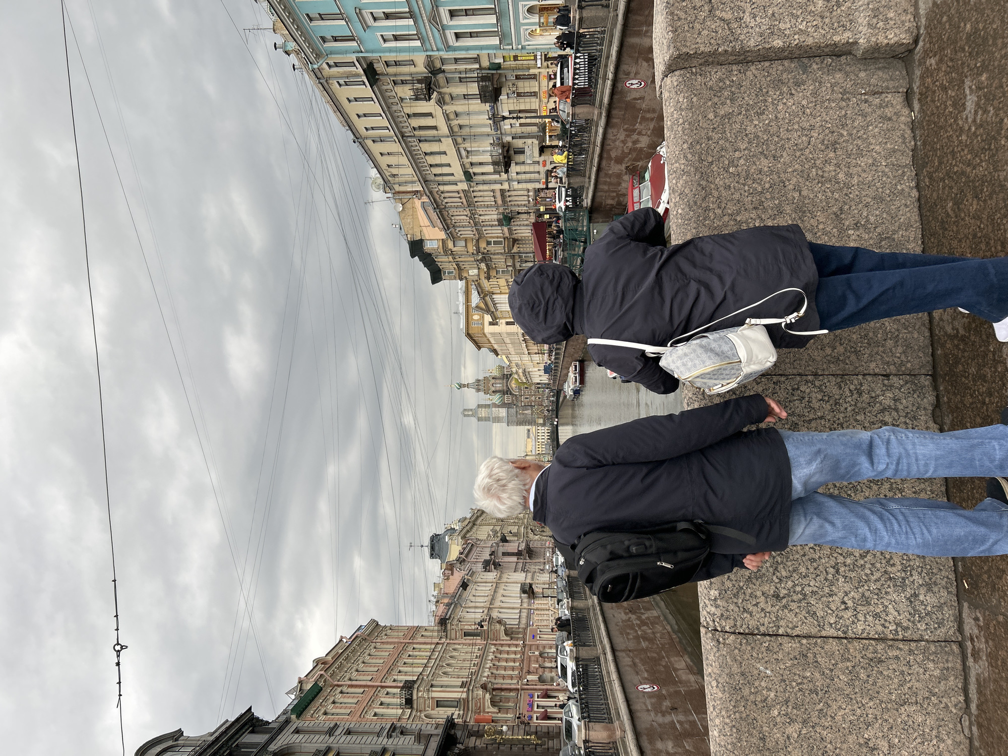 Two people are standing on a stone bridge looking over the edge into a canal in Saint Petersburg, Russia. The person on the right is wearing a dark hooded jacket, jeans, and white shoes. They have a small white purse on their shoulder. The person on the left has white hair and is wearing a dark jacket, jeans, and black shoes. They have a black backpack on their shoulder. Buildings line the canal on both sides and there are boats in the water. Power lines stretch across the sky. The sky is overcast and gray.