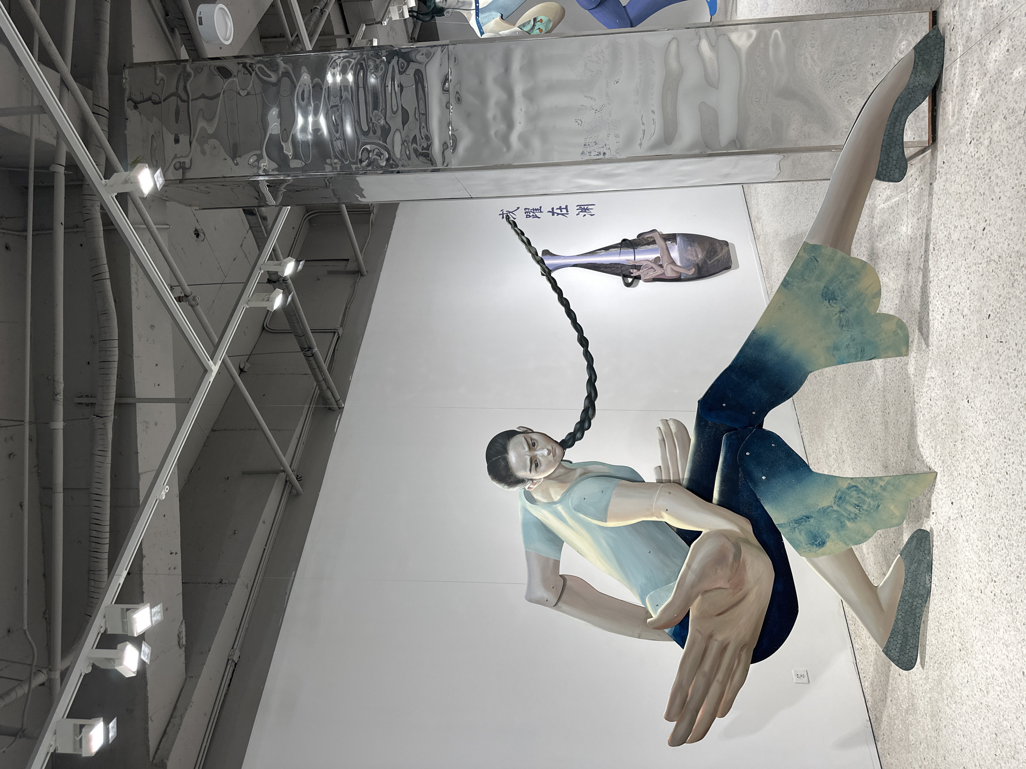 An indoor installation is displayed on a white wall. The exhibit appears to be a large cutout of a woman suspended by her hair. She is wearing a blue shirt and has large hands outstretched. Her legs are also cutout, with blue and green patterns on them. A silver fish-like object with a rope attached is mounted above her. The installation is located indoors, in what appears to be a gallery or exhibition space. The architecture and design of the space, including the ceiling and lighting, suggest a contemporary art environment. The location is in Banan, China.