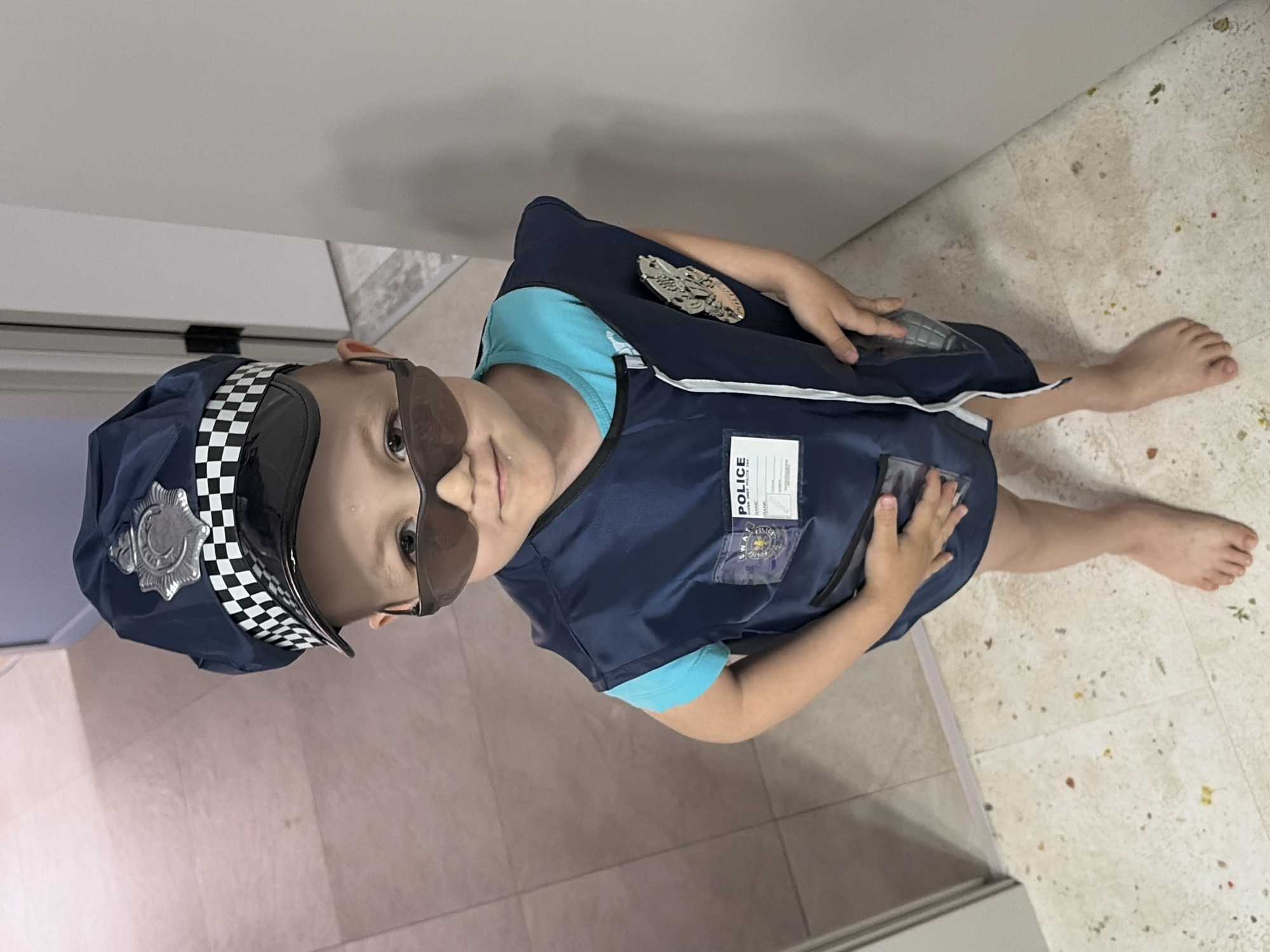 A child is dressed in a police costume. The child is wearing a police hat with a checkered band, sunglasses, and a navy blue vest over a light blue shirt. The vest has a police badge on the upper left side and a badge on the chest. The child is standing barefoot on a light-colored tile floor. The background includes a white wall and a portion of a doorway. The location is unknown.