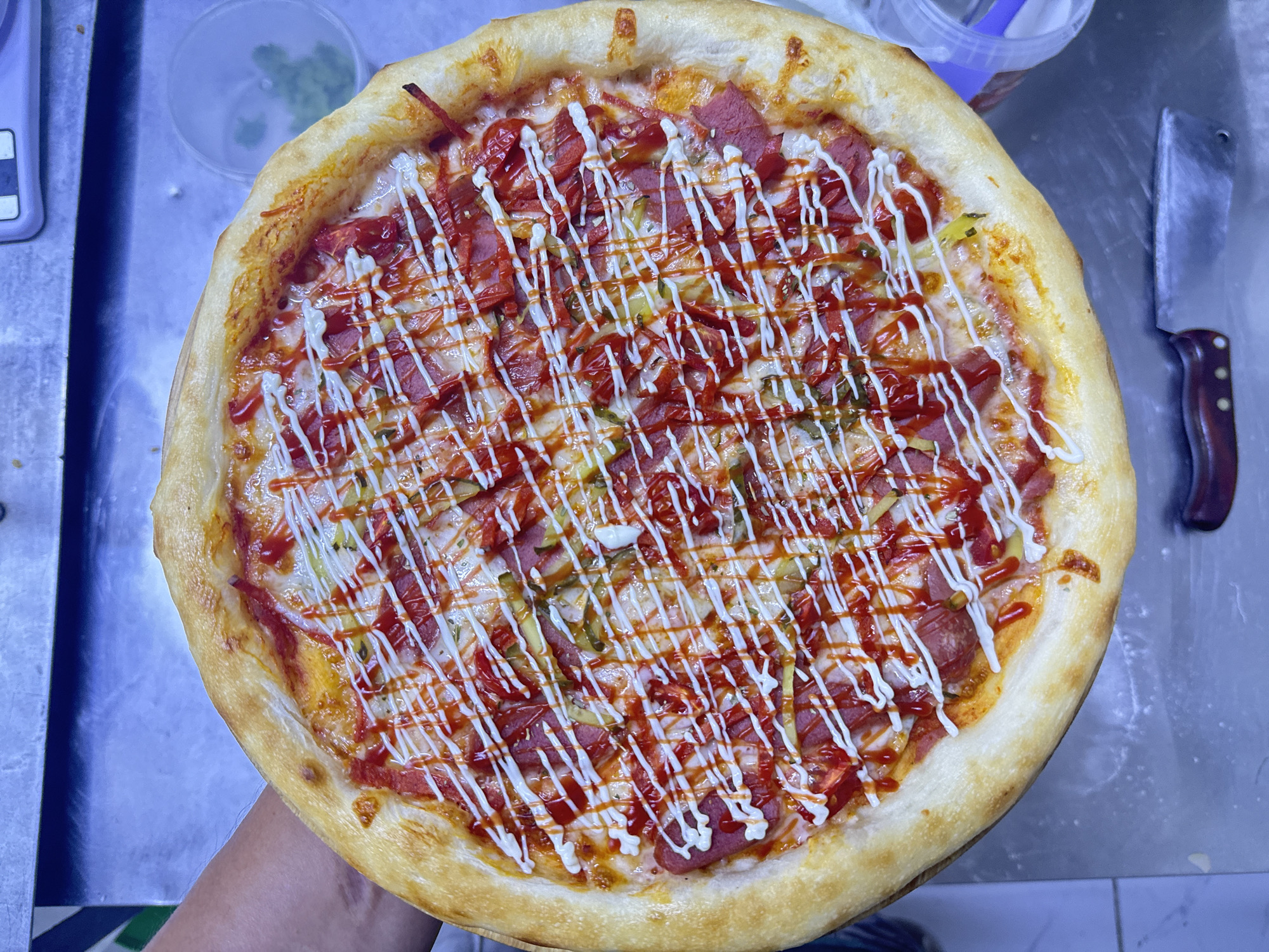 An overhead shot shows a person holding a whole pizza with a thick, golden-brown crust. The pizza is topped with a layer of melted cheese, sliced red sausage or pepperoni, and chopped green pickles. A drizzle of ketchup and mayonnaise crisscrosses the top of the pizza, adding a visually appealing pattern. The pizza appears to be fresh and ready to be served. The background is slightly blurred, suggesting the focus is entirely on the pizza. The environment appears to be a kitchen or food preparation area, with hints of metallic surfaces and a plastic container with green food items in the background. The location is identified as Ақши, Kazakhstan.