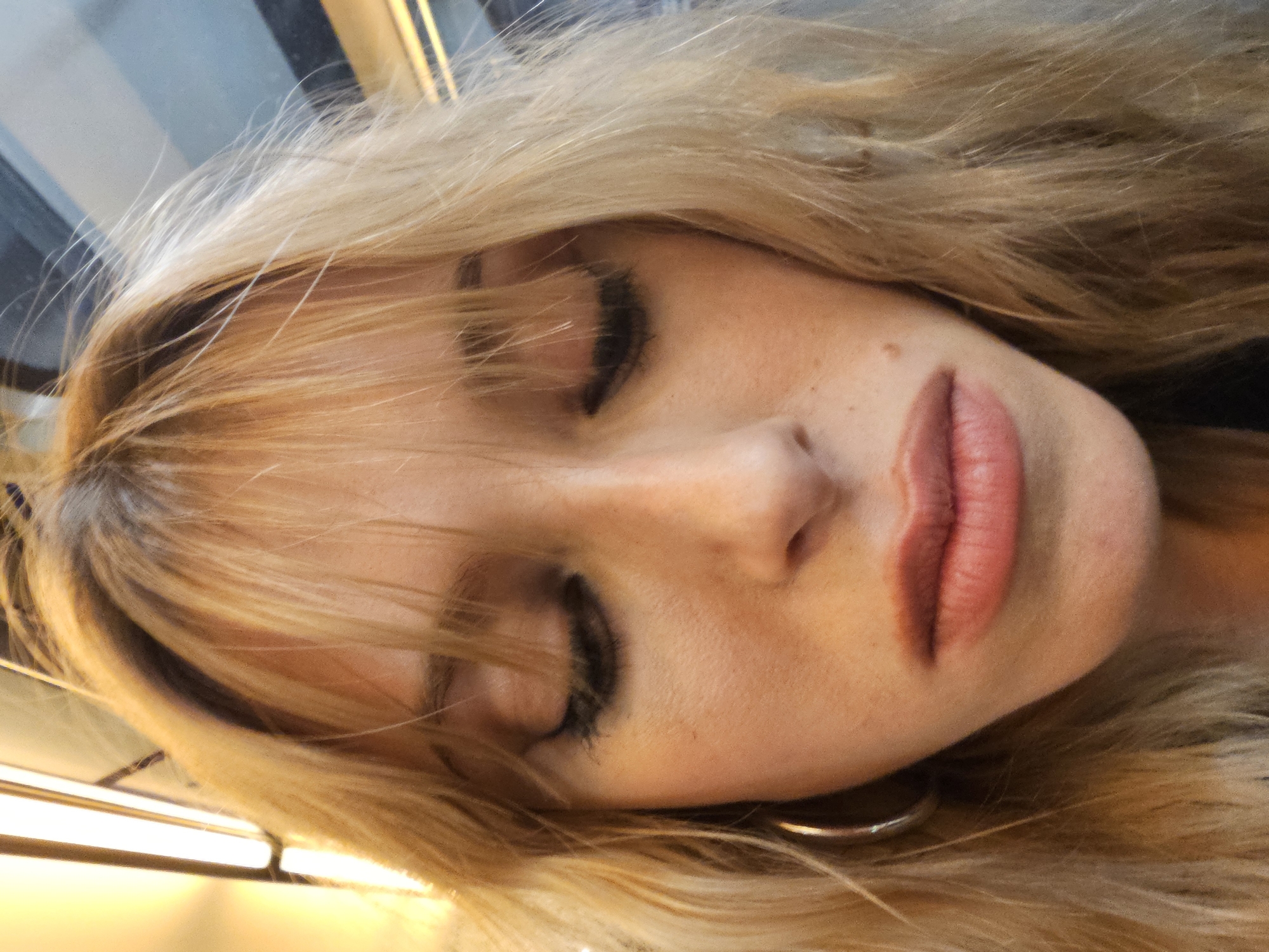 A close-up, high-angle shot captures a woman with blonde hair and closed eyes, seemingly asleep. Her face is tilted slightly to the side. She is wearing a gold hoop earring. The background is out of focus, but suggests a train or bus interior, possibly in Paris, indicated by the light reflections and indistinct city background seen through the window.