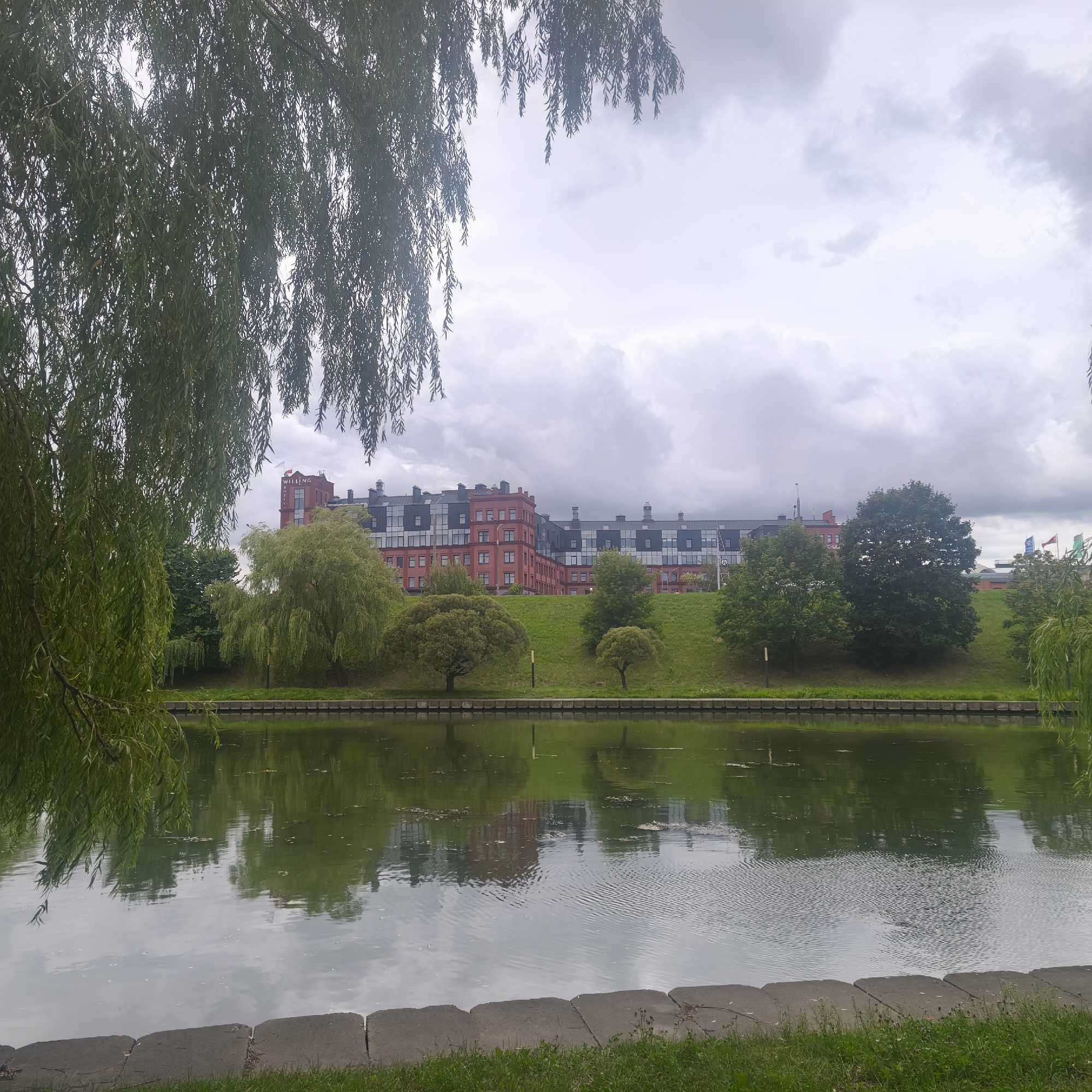 This image depicts a waterfront scene in Minsk, Belarus. A large, red brick building with dark-paneled sections and a white flag bearing the word "WILLING" is visible in the background, situated on a grassy hill. The building appears to be a hotel. In the foreground, a calm body of water reflects the sky and the surrounding greenery. Weeping willow trees hang over the water in the left foreground, and other trees are present on the grassy embankment. The sky is overcast with a thick layer of clouds. The scene is a tranquil, urban landscape.