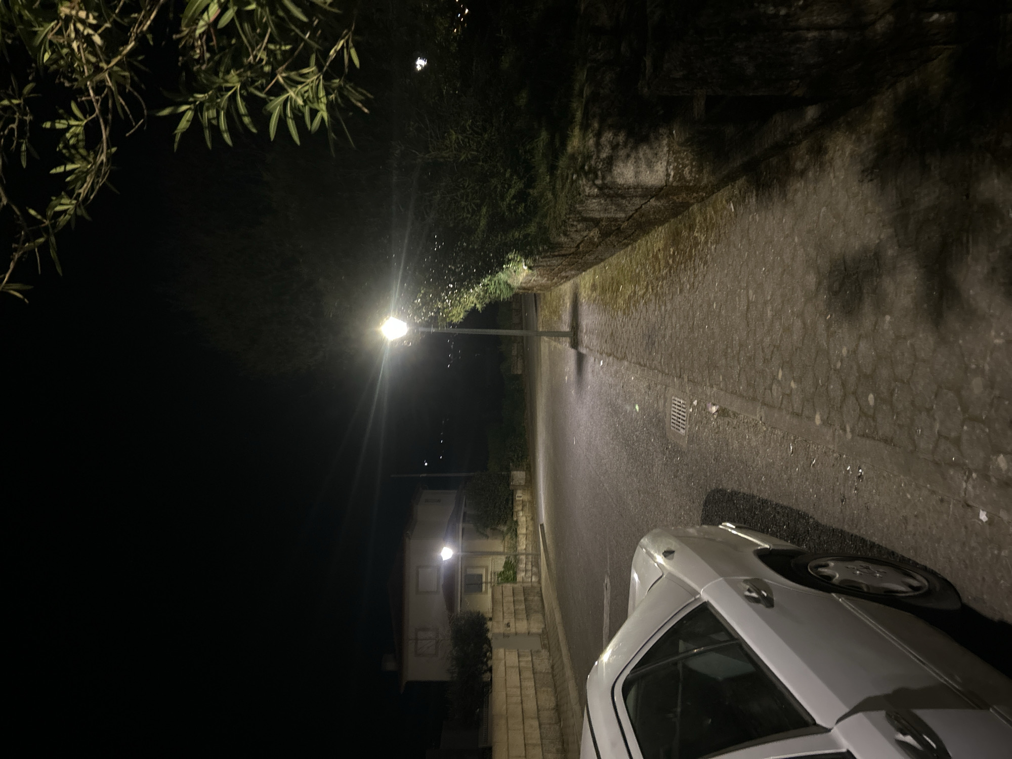 A nighttime view of a street in Vila Real, Portugal. The street is illuminated by a street lamp, with a car partially visible in the foreground. Buildings can be seen in the background, with trees and vegetation bordering the street. The pavement is visible and in good condition.