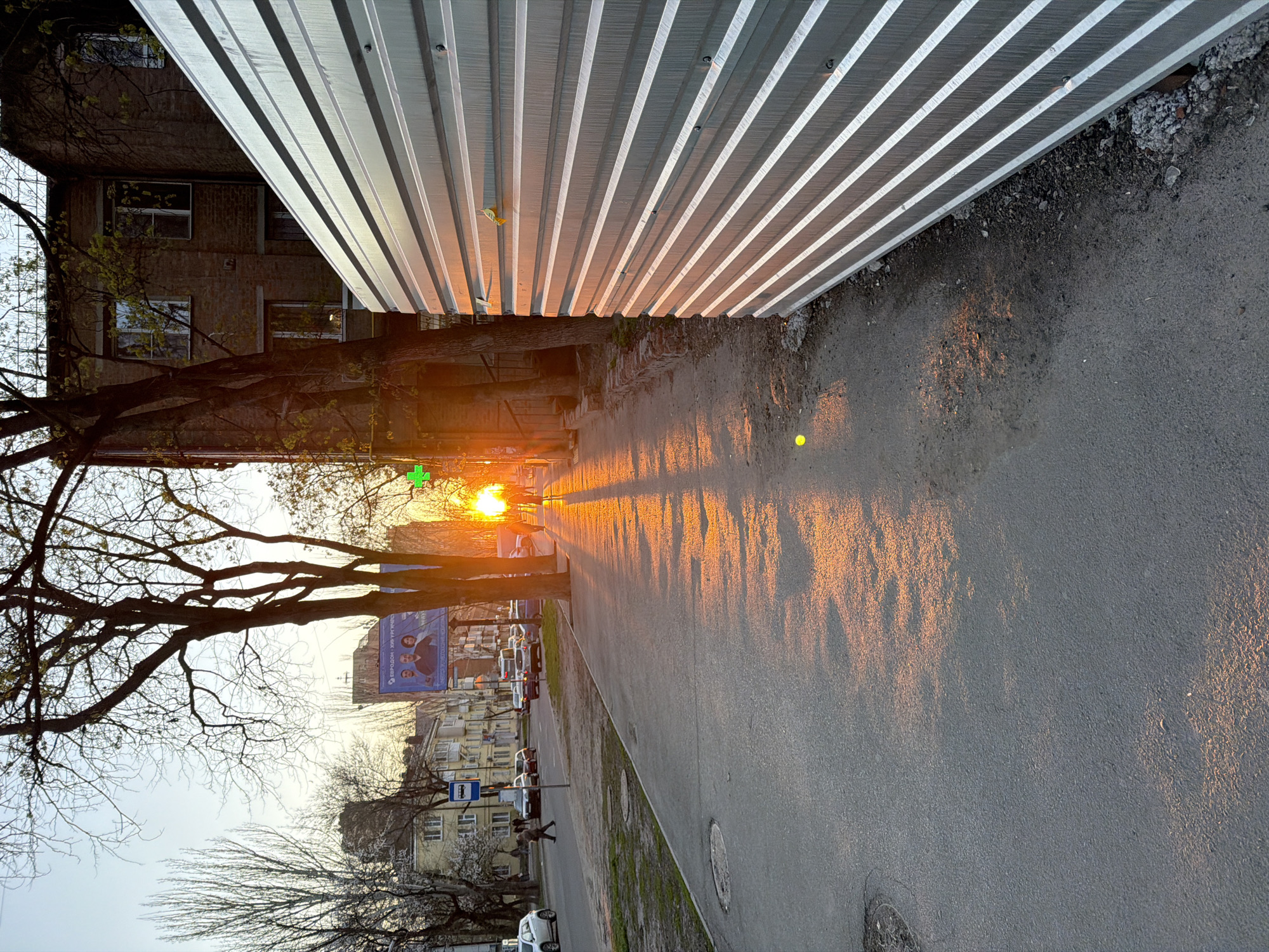 The image depicts a street scene in what is likely Rostov-on-Don, Russia. A metal fence runs along the top of the frame. A sidewalk and street run alongside it. The sidewalk and street are illuminated by bright sunlight, casting long shadows. Bare trees are visible, with a few nascent leaves. A bus stop sign and various parked cars can be seen further down the street. A building with a billboard is also visible in the distance. A tall building or apartment block is partially visible on the left.