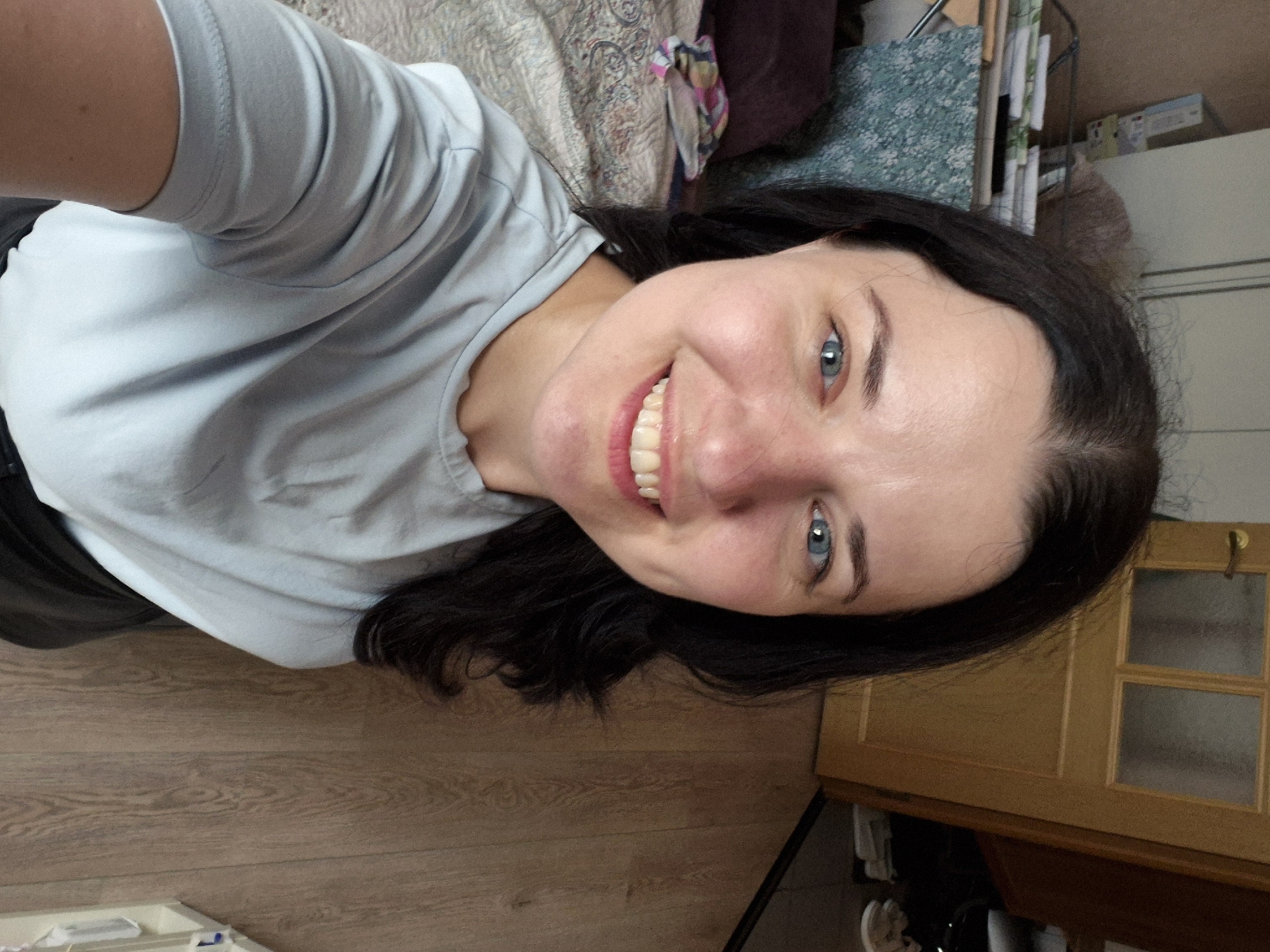 A woman with dark hair and blue eyes smiles at the camera. She is wearing a light blue t-shirt and is positioned in what appears to be a bedroom, with a bed to her left and a wooden floor and door to her right. The scene is indoors, likely in an apartment or house.