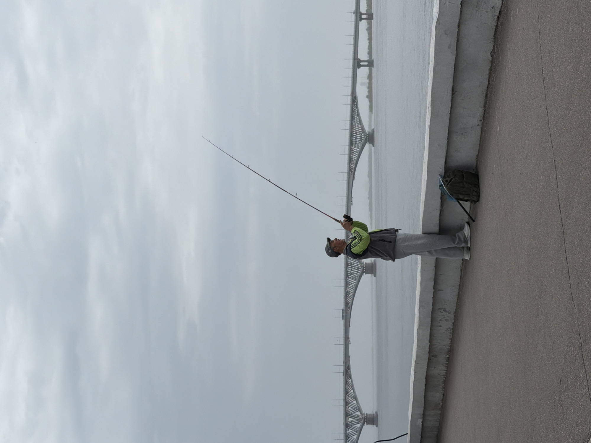 A man is fishing from an elevated walkway near a river in Saratov, Russia. The angler wears light grey sweatpants and tennis shoes, a light grey jacket, and a dark baseball cap. He holds a long fishing rod in his hands, angling it out over the water. Next to the man, on the walkway, rests a green backpack. Behind him and across the river is a bridge that has a web-like design. The sky is overcast.