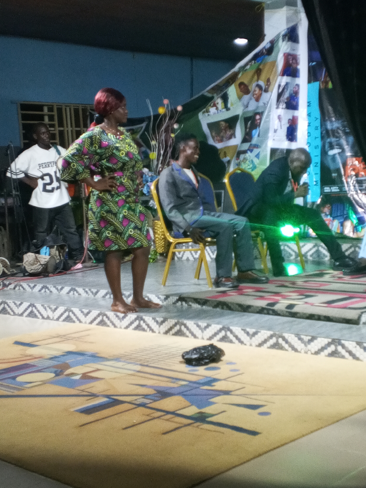 A medium shot indoors captures a scene on a slightly elevated stage. The location is presumed to be in the city of Unknown, Unknown. On the stage, three people are present. A woman in a vibrant green, black, and pink patterned dress stands barefoot on the left, her hands on her hips, and her red hair is a notable feature. To her right sits a man in a grey suit, smiling slightly. Another man in a dark suit is seated to the right of the first man, seemingly engrossed in an activity involving his hands, and a soft green light emanates near his legs.
Behind the seated men is a large banner with multiple photos and the words "ODRAM MINISTRY" printed on it. To the left of the woman, a man in a "PERRYPA" jersey with the number 25 is standing. Various objects, including a bag and a coiled wire, are on the stage near him.
The stage is decorated with a patterned rug, and a dark object is situated in the foreground, on a beige and blue rug. The background is a muted blue, with a window visible behind the standing man.