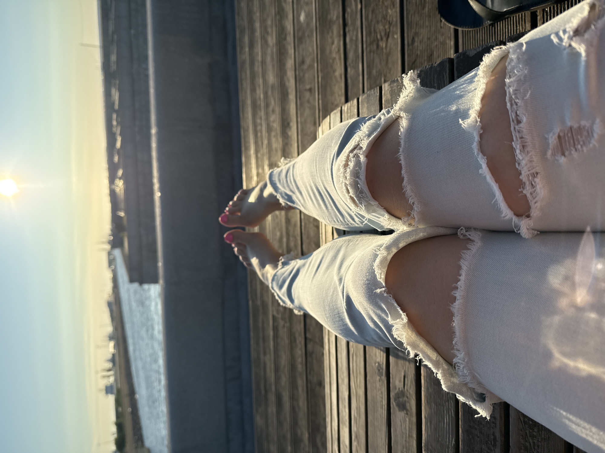 This is a close-up, low-angle shot of a person's legs, wearing light-wash ripped jeans and nail polish on their toes, resting on a wooden deck. The sun is setting or rising, casting a warm glow and creating lens flare. In the background, a body of water with boats and a hazy cityscape is visible. The location is likely a waterfront area in Saint Petersburg, Russia, possibly near a harbor or embankment, given the presence of boats and the characteristic architecture suggested by the distant skyline. The scene captures a moment of relaxation in an urban outdoor setting.