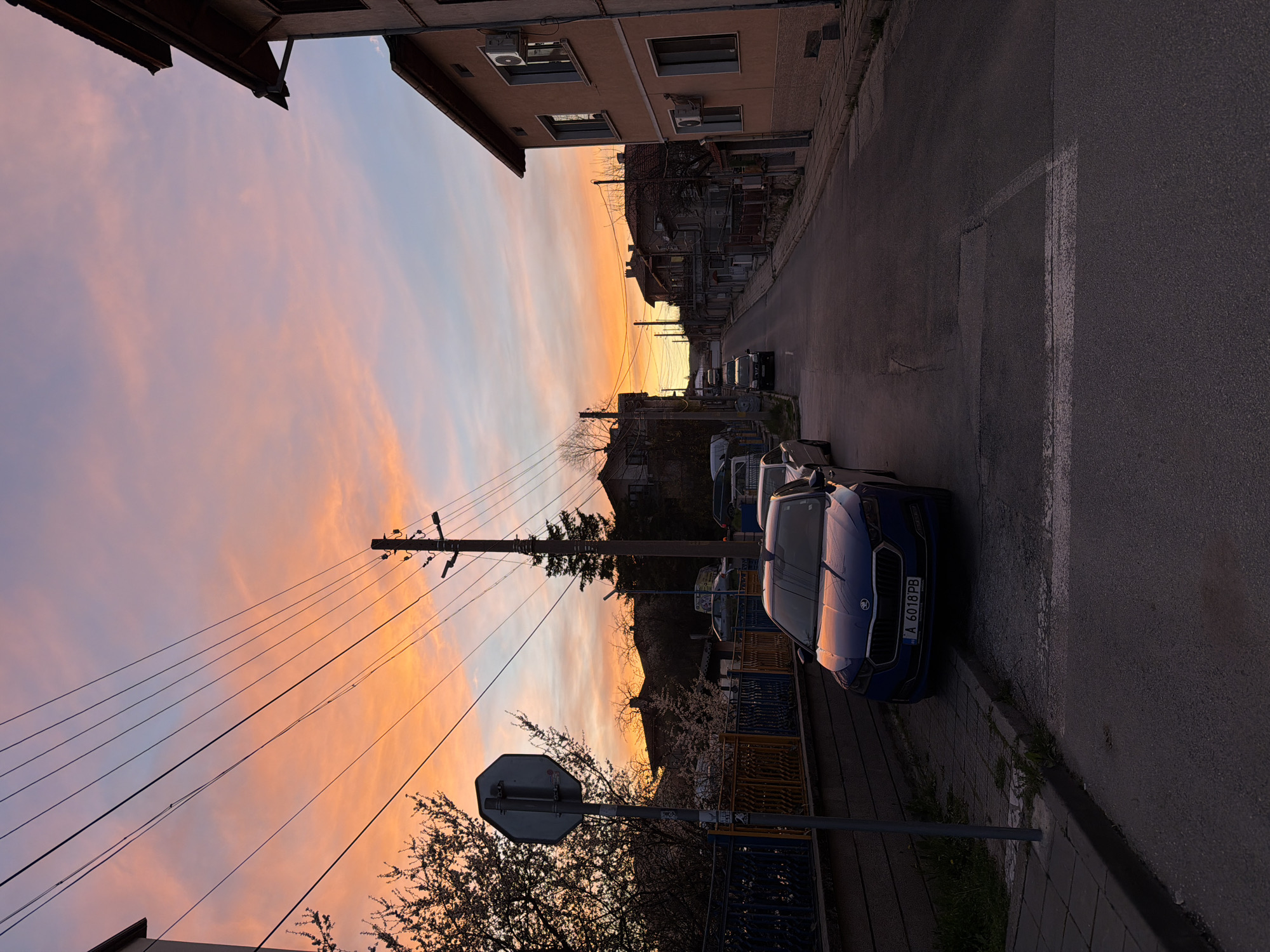 The photograph captures a street scene in Panagyurishte, Bulgaria, during sunset. The sky is a vibrant gradient of pink, orange, and blue hues, with wispy clouds scattered across the expanse. The street is lined with buildings, one of which appears to be a multi-story apartment block with air conditioning units visible on its facade. Parked along the curb are several cars, including a blue sedan in the foreground with license plate "A 6018 PB". Utility poles stretch along the street, with power lines crisscrossing the sky. A street sign is visible near the curb. The scene is cast in a combination of warm sunset light and shadow.