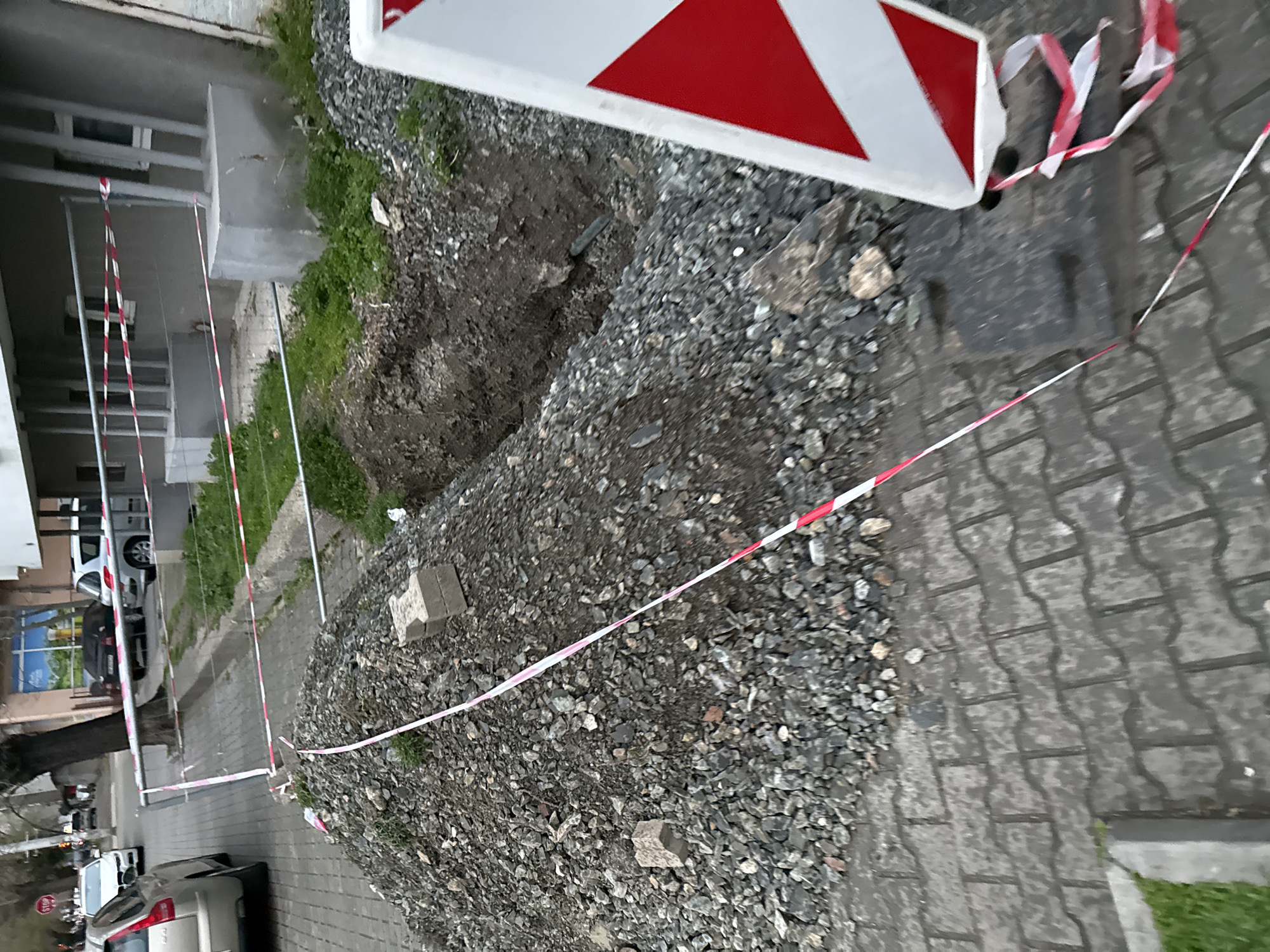 The image depicts a construction site on a paved street in Stara Zagora, Bulgaria. A pile of gravel and dirt is visible, cordoned off by red and white striped caution tape. A red and white striped road sign is partially visible on the right side of the image, adjacent to the gravel pile. In the background, buildings and parked cars are visible.