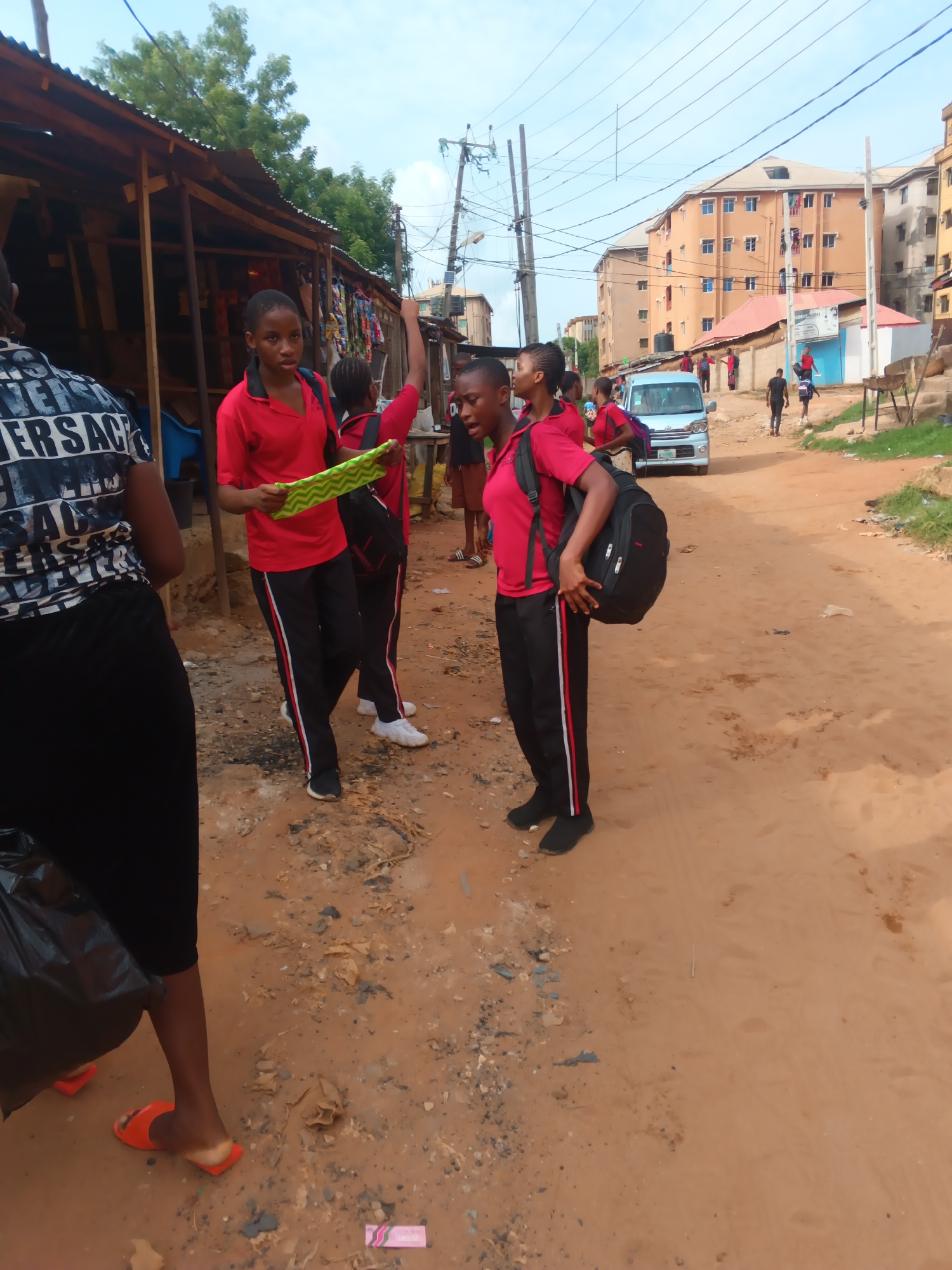 The image shows a group of students in red shirts and black pants with red and white stripes, likely school uniforms, standing on a dirt road in Onitsha, Nigeria. One student is holding a green and yellow object. Another student wears a backpack. There are wooden structures, possibly market stalls, on the left. A white minivan and several other people are in the background. Buildings and power lines are visible in the distance. The ground is dirt and gravel. A person wearing a "Versace" shirt, orange sandals, and a black bag is on the left of the scene. A pink wrapper lays on the ground.